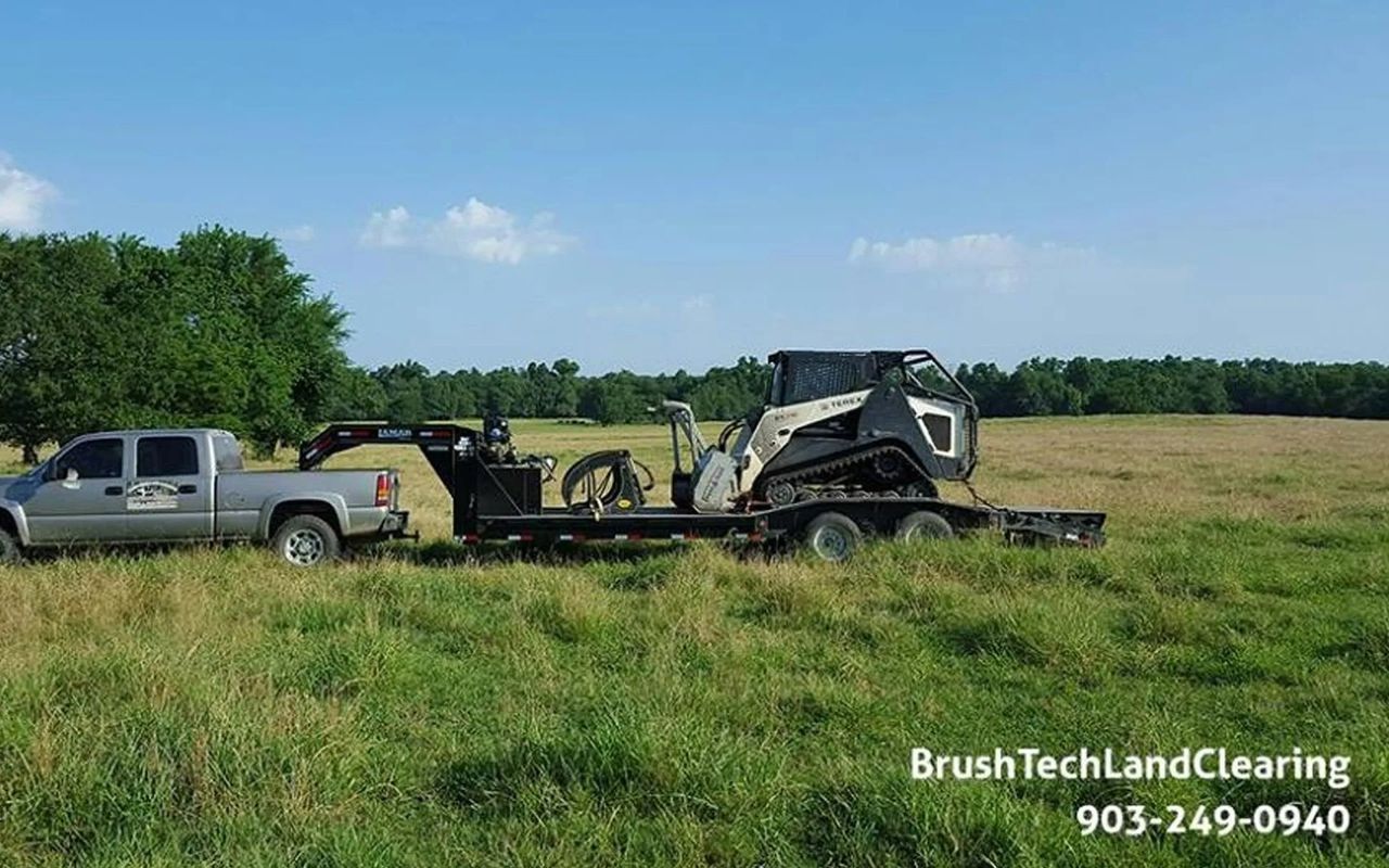 A truck towing a trailer with a skid steer on a grassy field under a blue sky.
