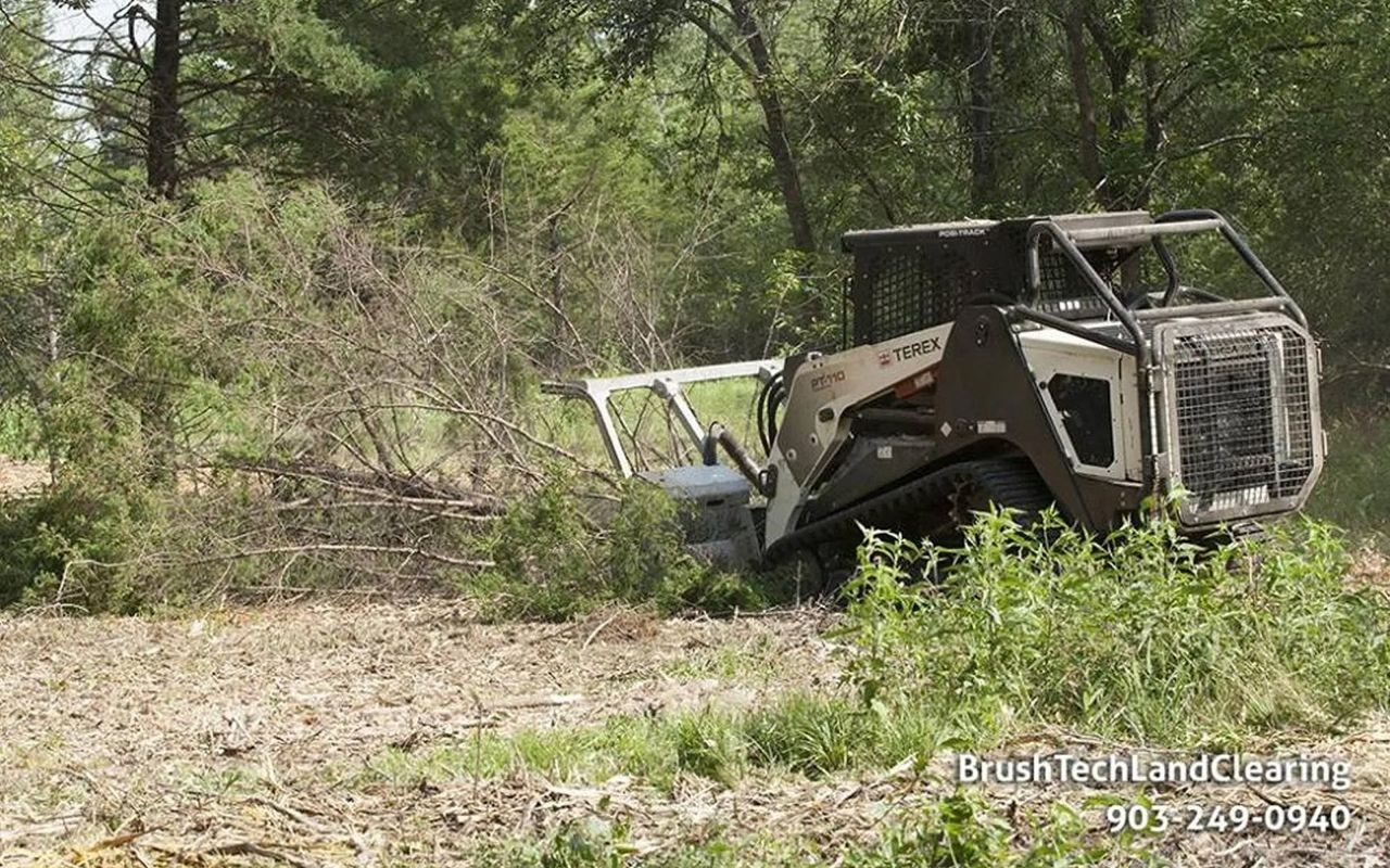 Bobcat clearing brush in a field. White machine with open door. Green trees in background.