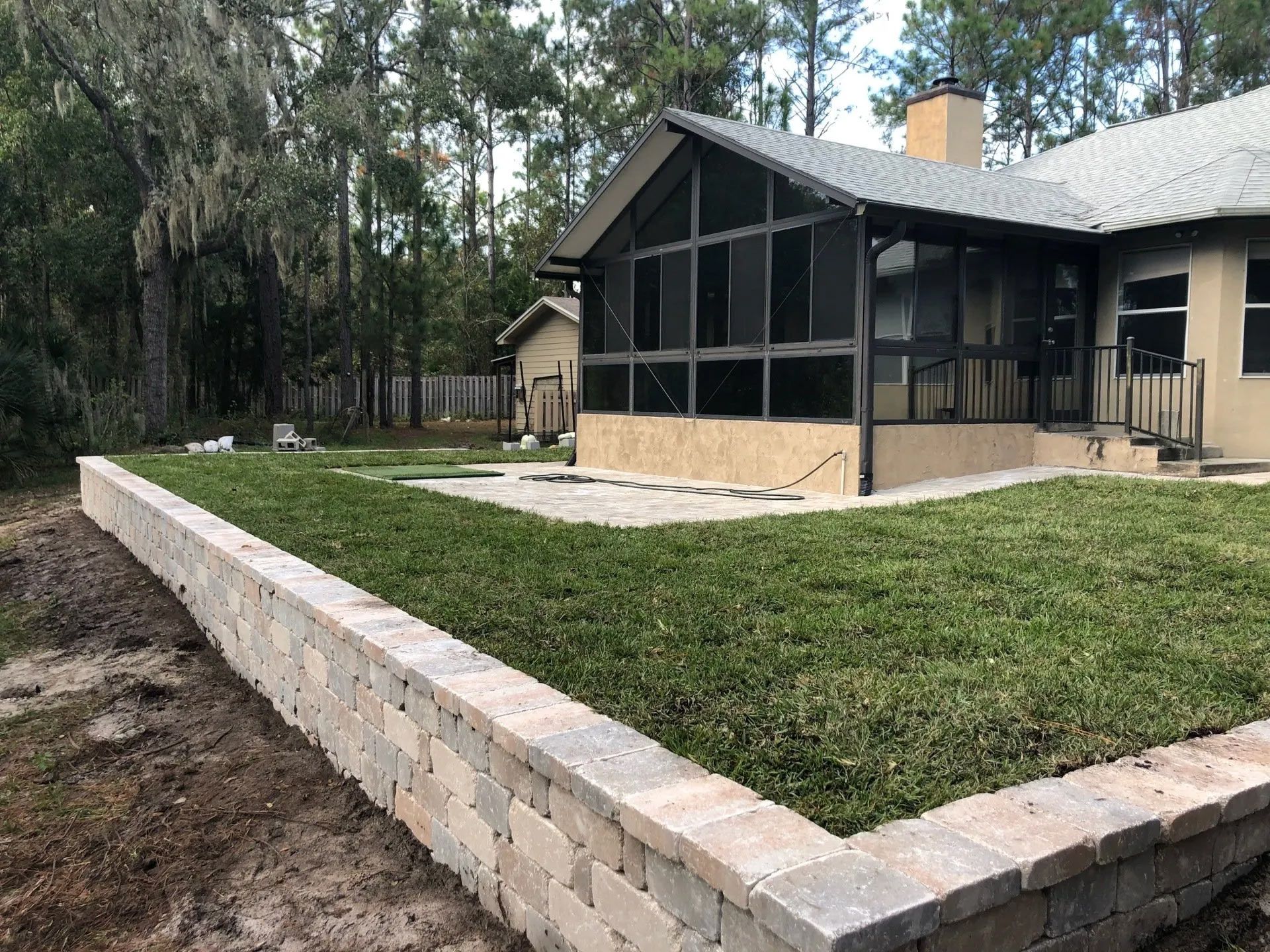 A retaining wall of tan and gray blocks borders a grassy yard next to a house with a screened porch.