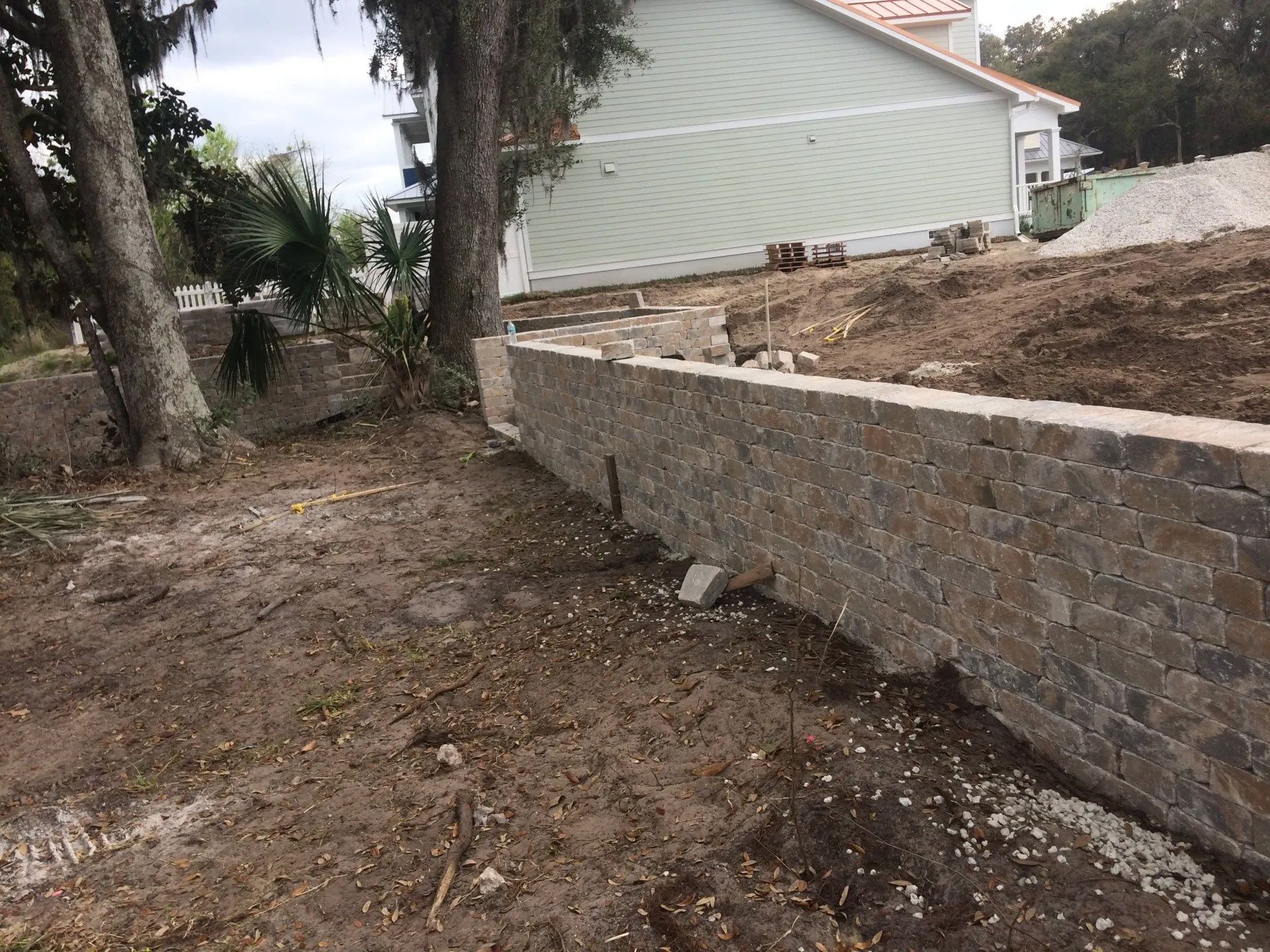 A retaining wall made of stacked brown bricks in front of a light green house under construction. The ground is dirt.