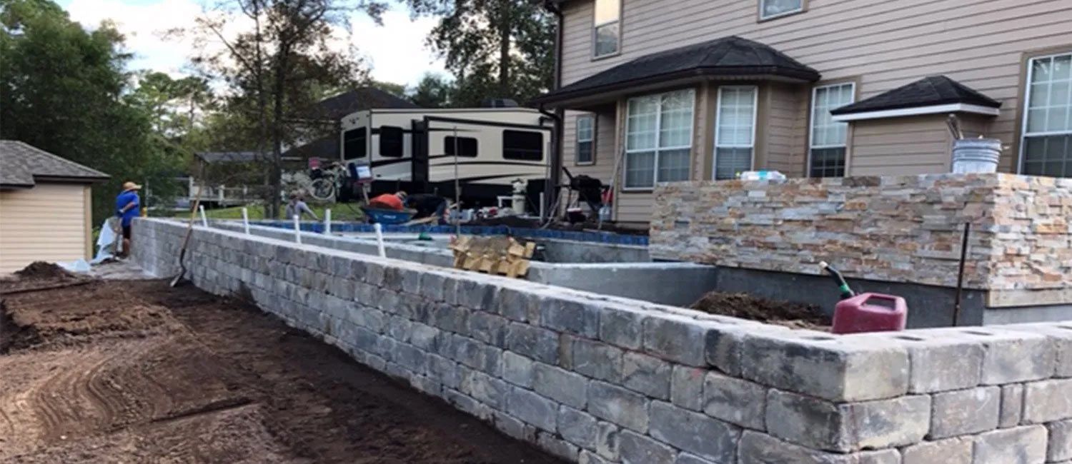 Construction site with a brick retaining wall in front of a house. A motorhome and workers are visible in the background.