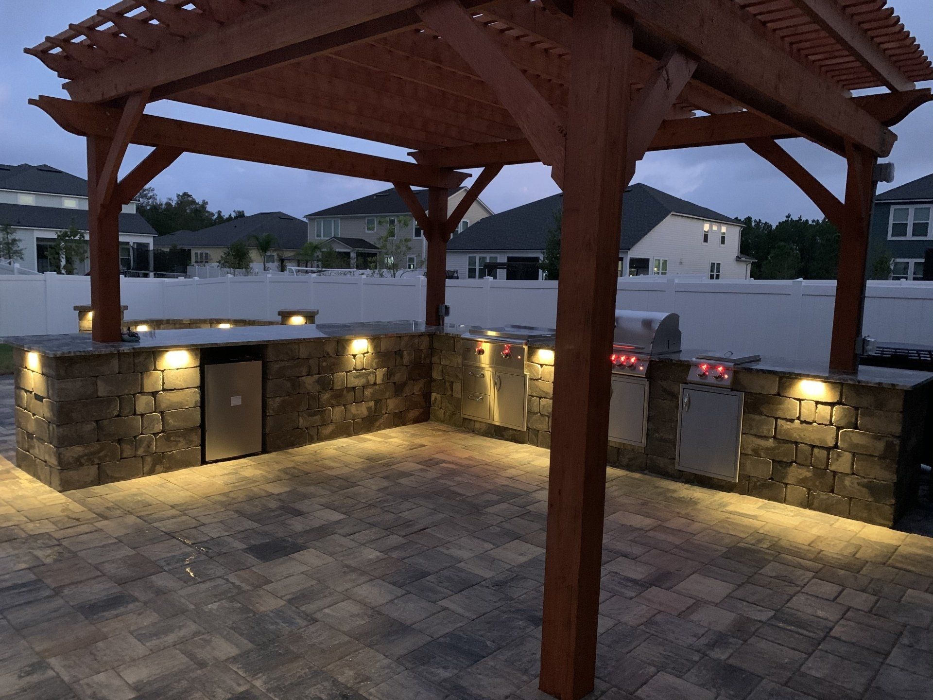 Outdoor kitchen under a wooden pergola with stone facade, appliances, and built-in lighting. Paved patio with houses in the background.