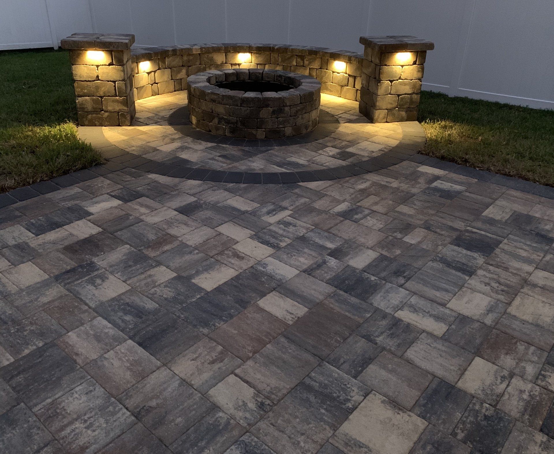 A brick patio with a fire pit, accented by warm lighting on stone pillars and surrounding wall, in a yard.