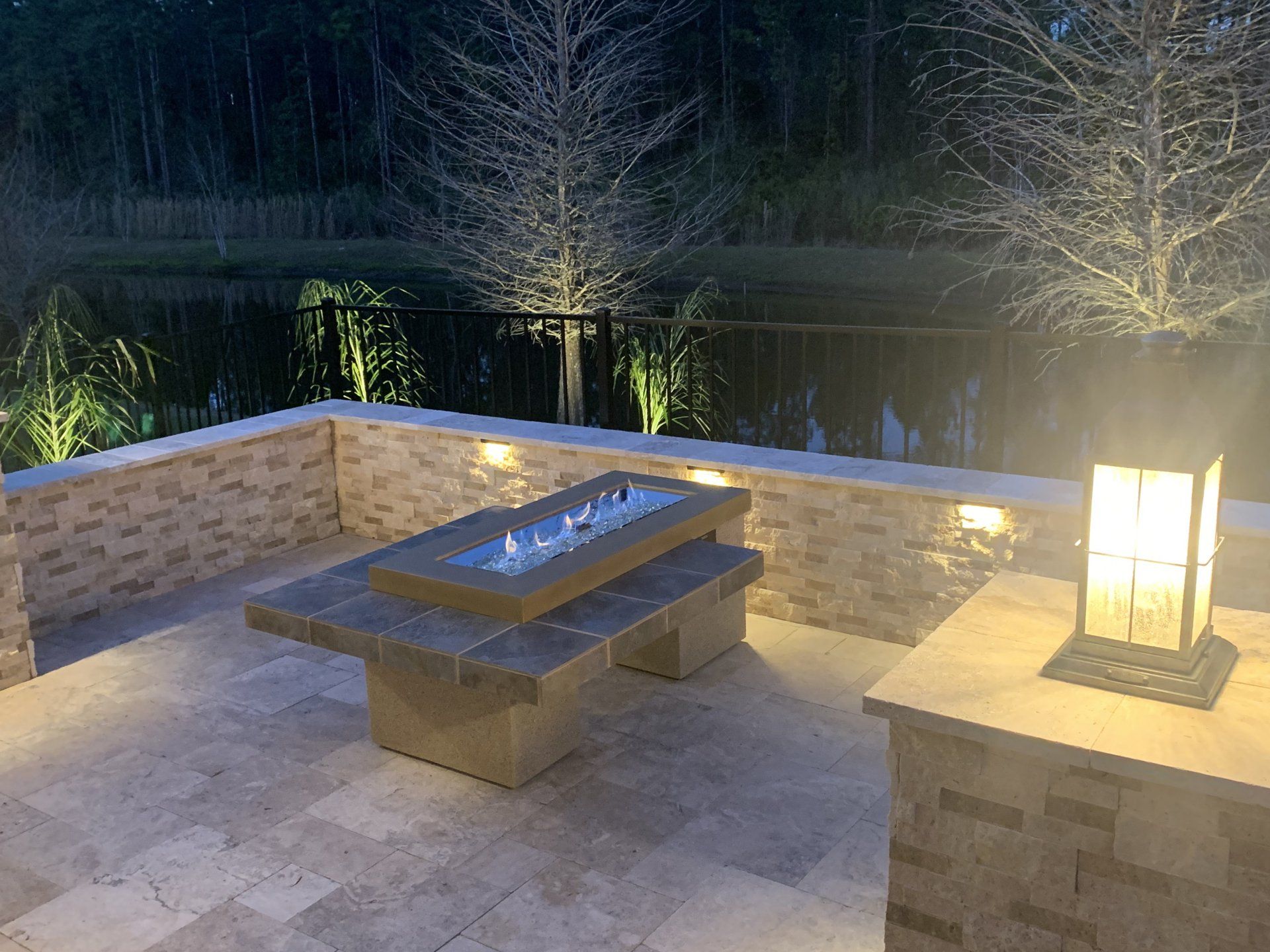 Outdoor patio at dusk with a fire pit, seating, and accent lighting overlooking a pond. Brickwork and neutral tones dominate the scene.