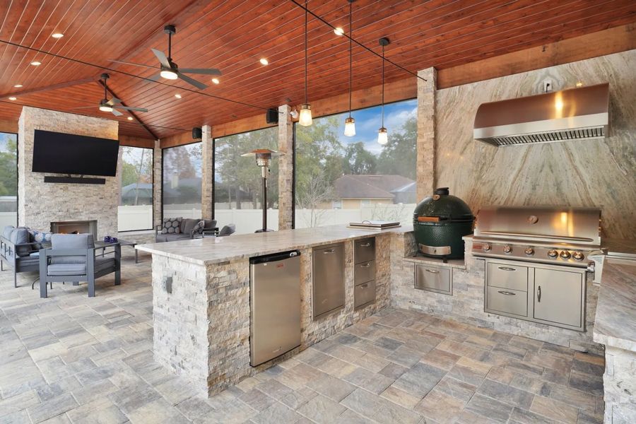 Outdoor kitchen with a stone island, grill, and Big Green Egg. Wooden ceiling with fans and recessed lighting, view of a backyard.