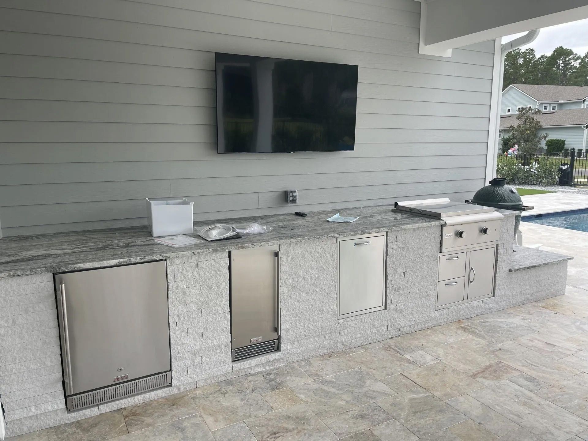 Outdoor kitchen with granite countertop, stainless steel appliances, and a mounted TV against a gray siding wall; a swimming pool is visible in the background.