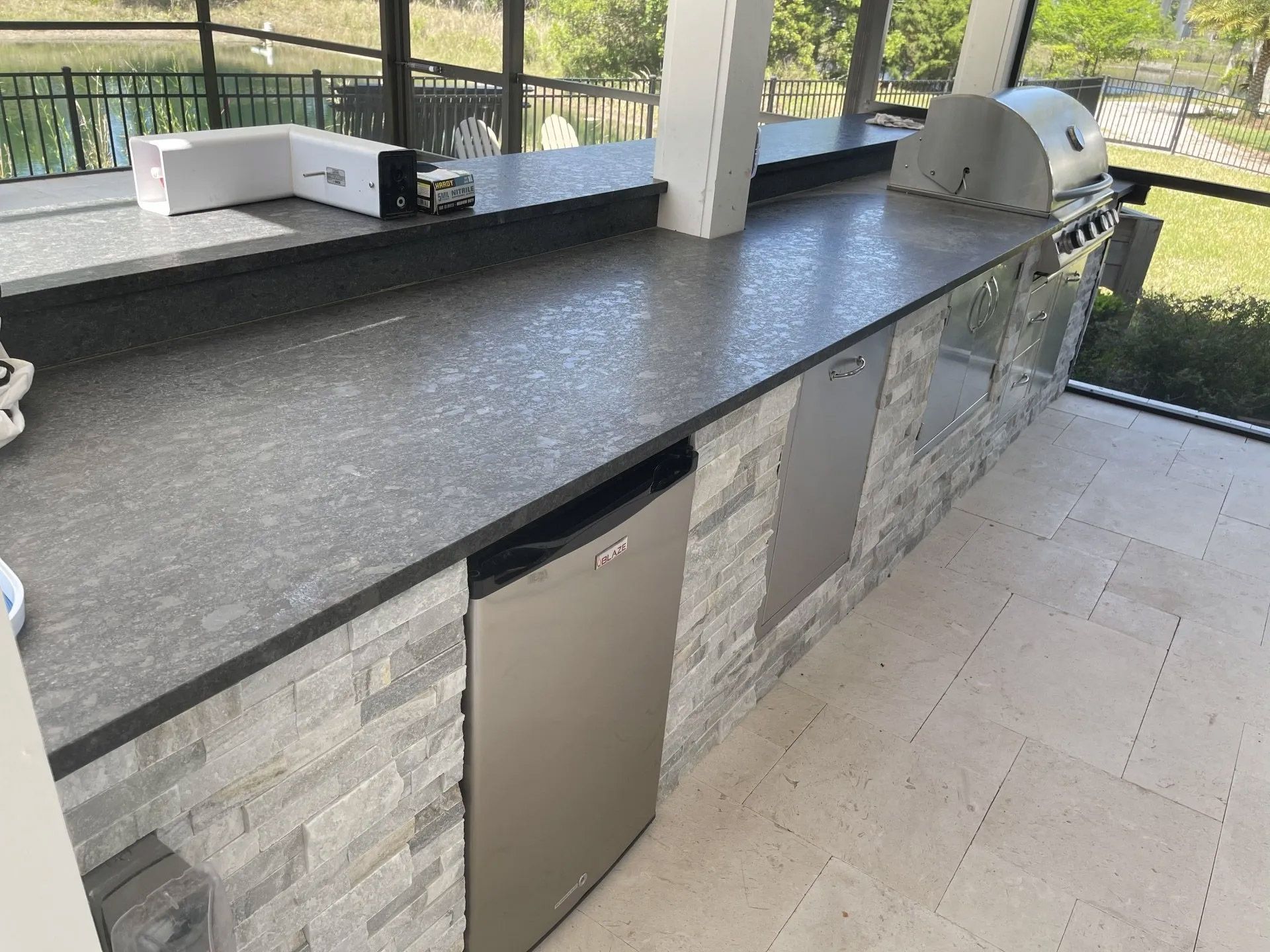 Outdoor kitchen with gray countertop, stacked stone facade, stainless steel appliances including a refrigerator and grill.