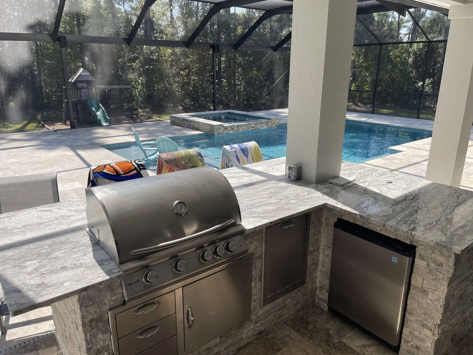 Outdoor kitchen with a stainless steel grill, and a pool in the background. The counter is granite with a dishwasher and cabinet.