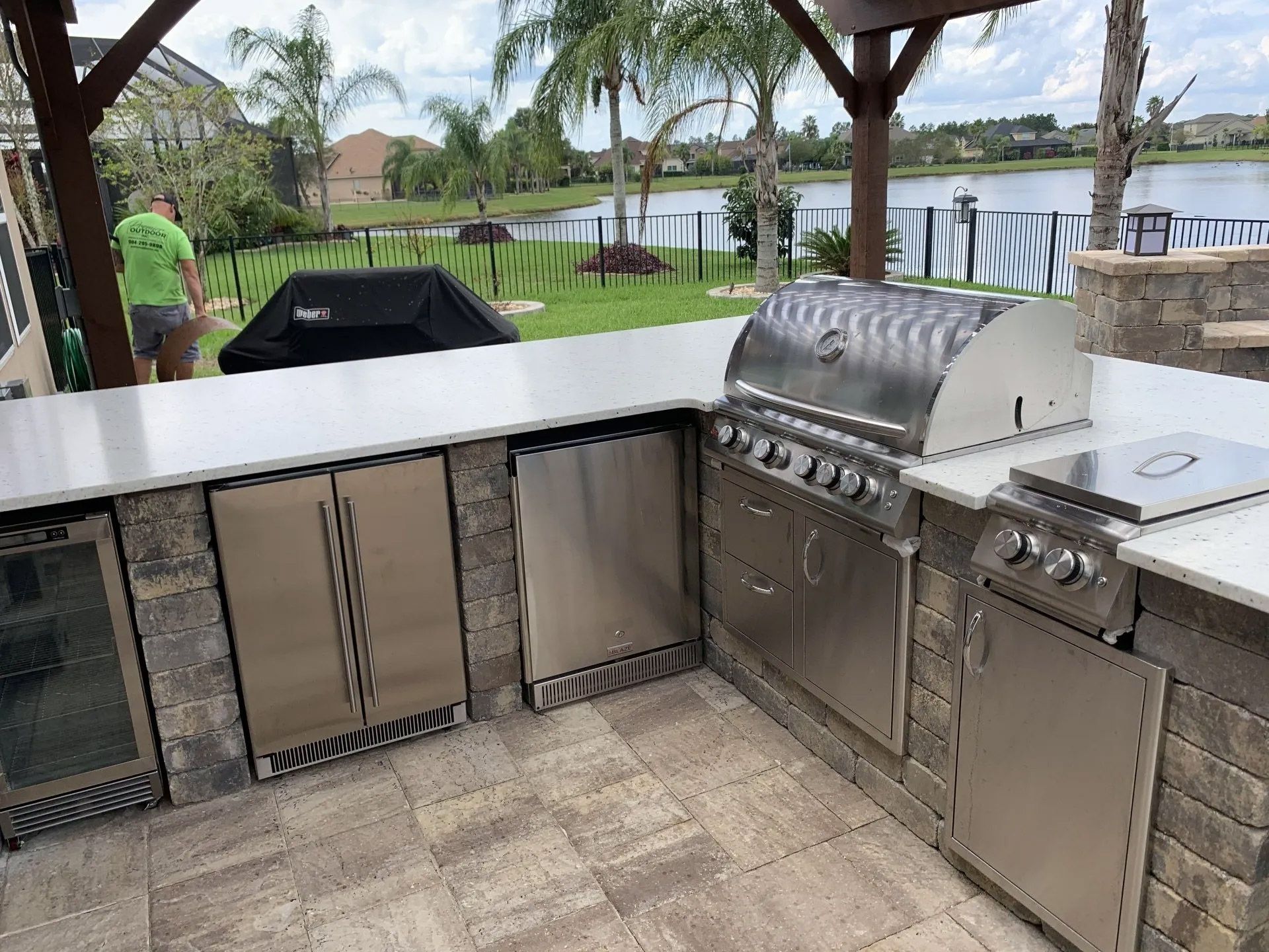 Outdoor kitchen with a stainless steel grill, two refrigerators, and a person in a green shirt in the background near a lake.