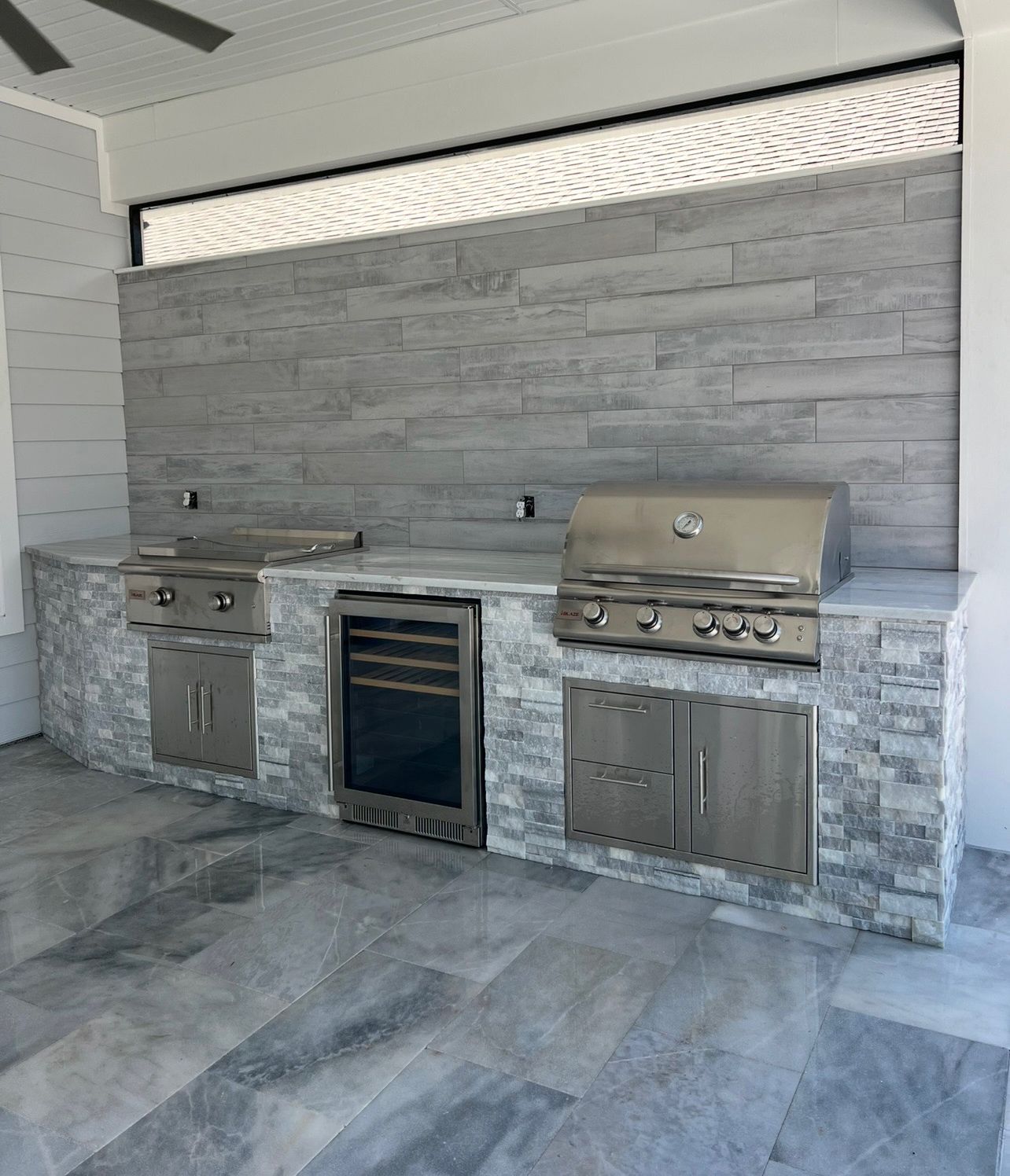 Outdoor kitchen with stainless steel grill, cooktop, and refrigerator built into a gray stone counter and backsplash.