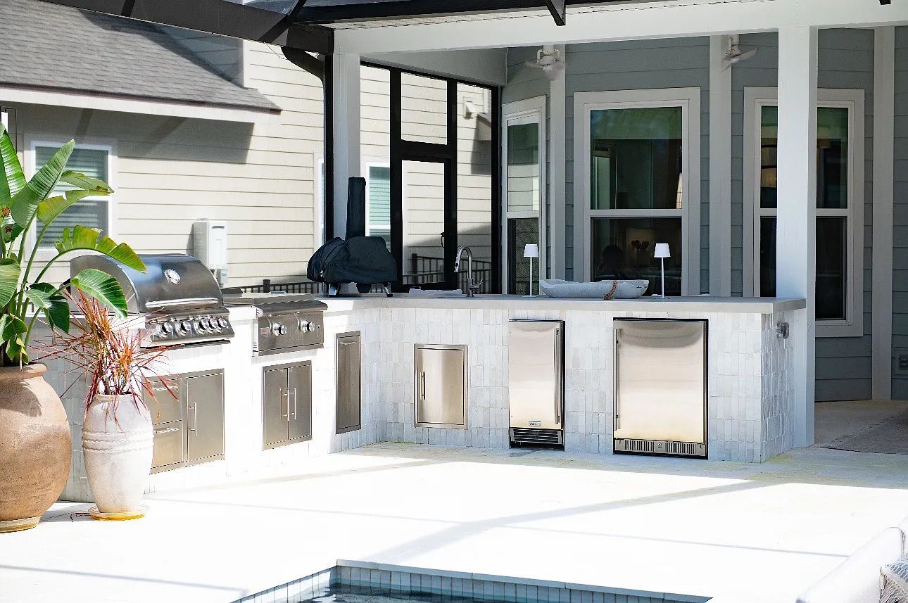 Outdoor kitchen with grill, refrigerator, and sink under a covered patio. Stainless steel appliances contrast with light gray cabinetry.