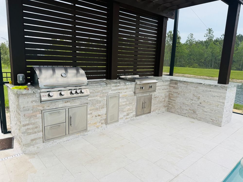Outdoor kitchen with a grill, cabinets, and built-in appliances, under a dark slatted pergola, near a pool.