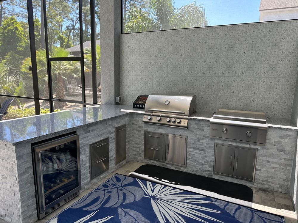 Outdoor kitchen with a stainless steel grill, refrigerator, and cabinets, set against a light gray stone wall and a blue and white rug.