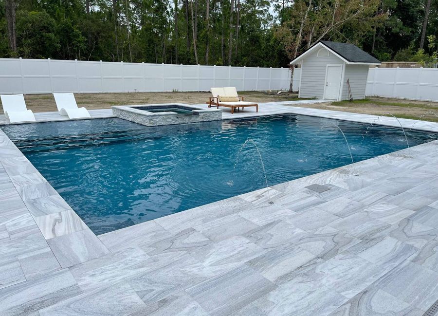 A rectangular pool and spa with light gray stone surround, two lounge chairs, and a small white shed.