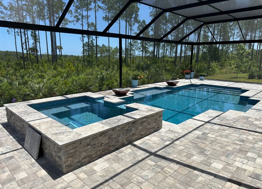 A screened-in outdoor pool with a hot tub, surrounded by stone pavers, and overlooking a wooded area on a sunny day.