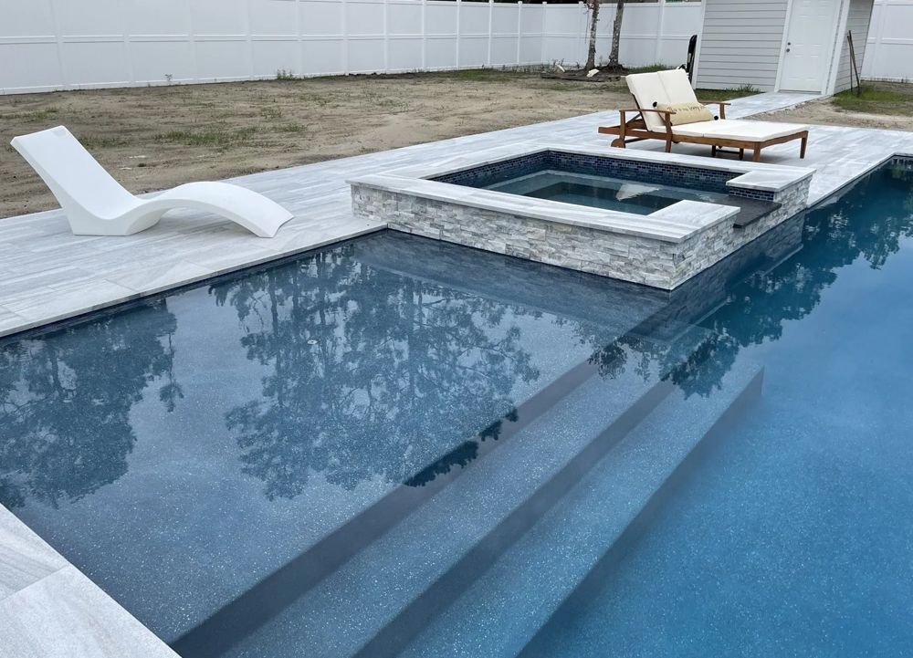 A rectangular swimming pool with a built-in hot tub, steps, and a lounge chair, with a white fence in the background.