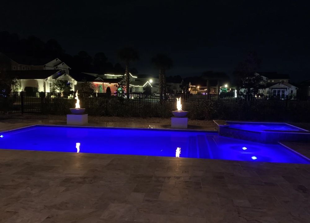 Night view of a backyard pool with blue underwater lights, two fire features, and illuminated houses in the background.