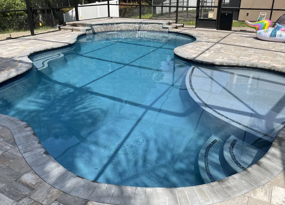 A blue swimming pool with stone coping and a built-in spa, surrounded by a tiled patio, viewed from above.