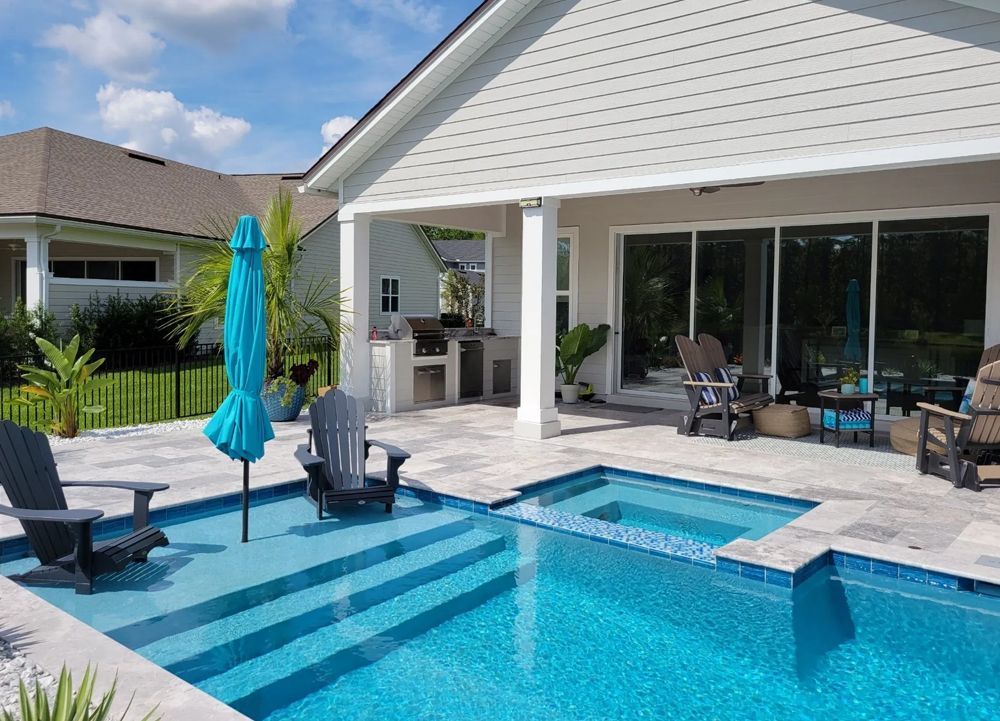 A backyard with a pool and patio. Blue pool water, adirondack chairs, and umbrella, next to a covered seating area with a grill.