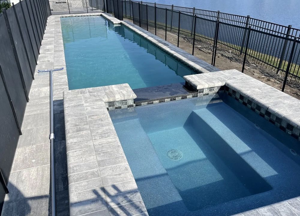 A rectangular swimming pool with a connected spa, both filled with water. The pool area is surrounded by gray stone and a black fence.