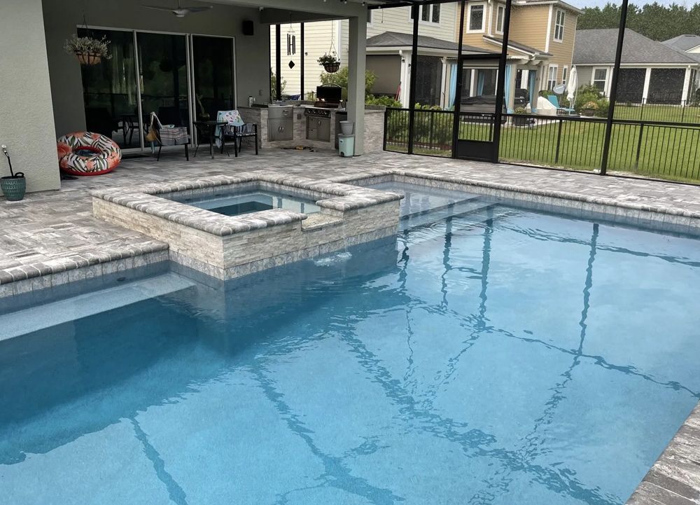 A swimming pool with attached hot tub, featuring blue water and a stone patio. In the background is a screened-in patio and residential homes.