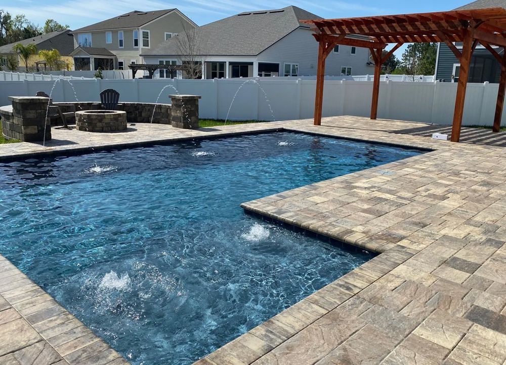Backyard pool with blue water, fountains, and a wooden pergola, surrounded by paved patio and a fire pit.