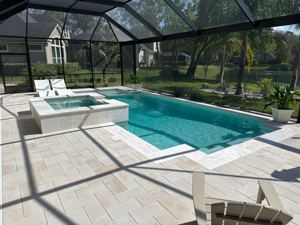 A screened-in pool area with a rectangular pool, hot tub, and light-colored stone patio. There are two lounge chairs and a scenic backyard view.