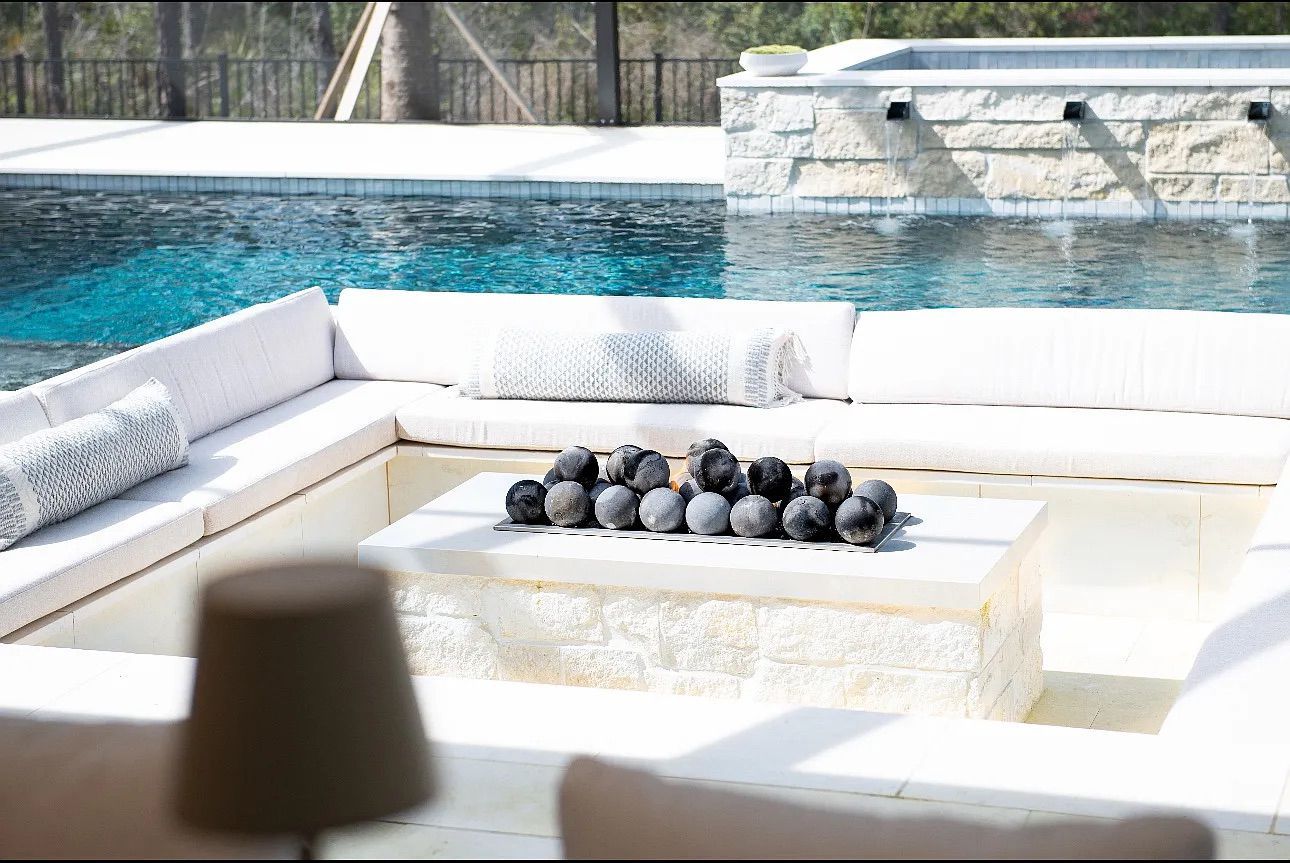 Outdoor seating area with a fire pit and pool. White cushions and stone surround the fire pit and pool.