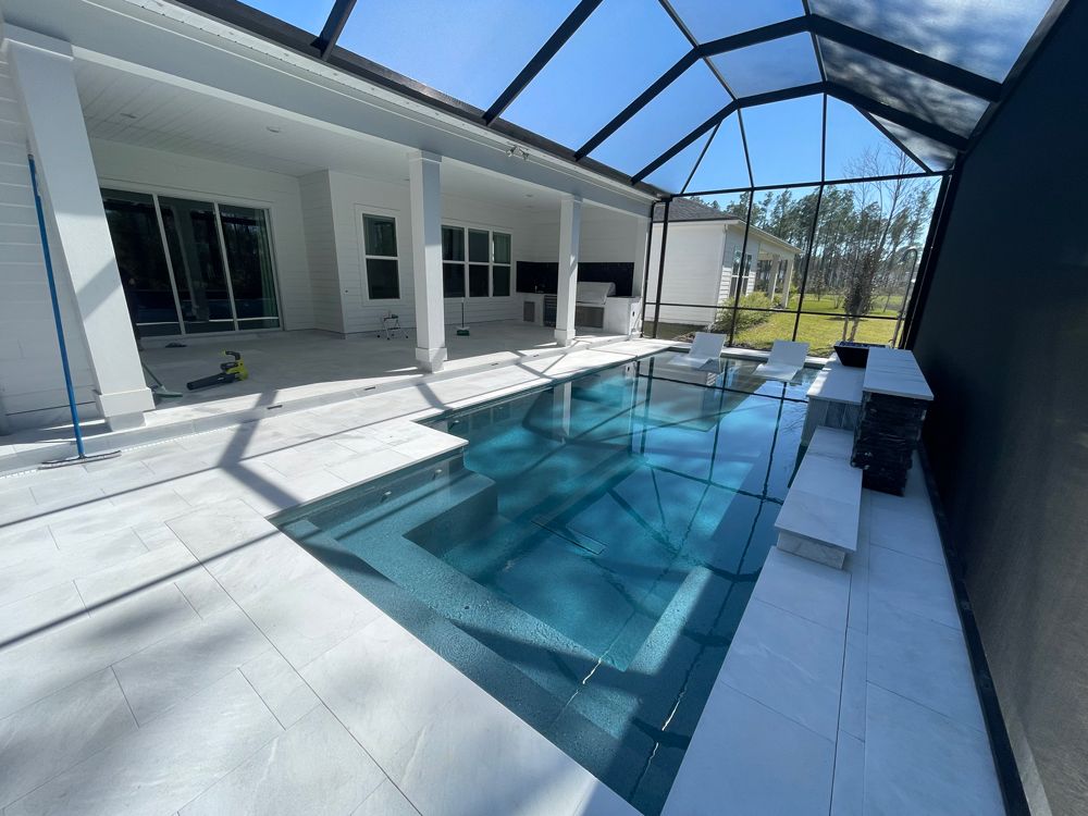 A luxurious, modern pool with a dark-blue tiled interior, surrounded by a white-tiled patio and screened enclosure, on a sunny day.