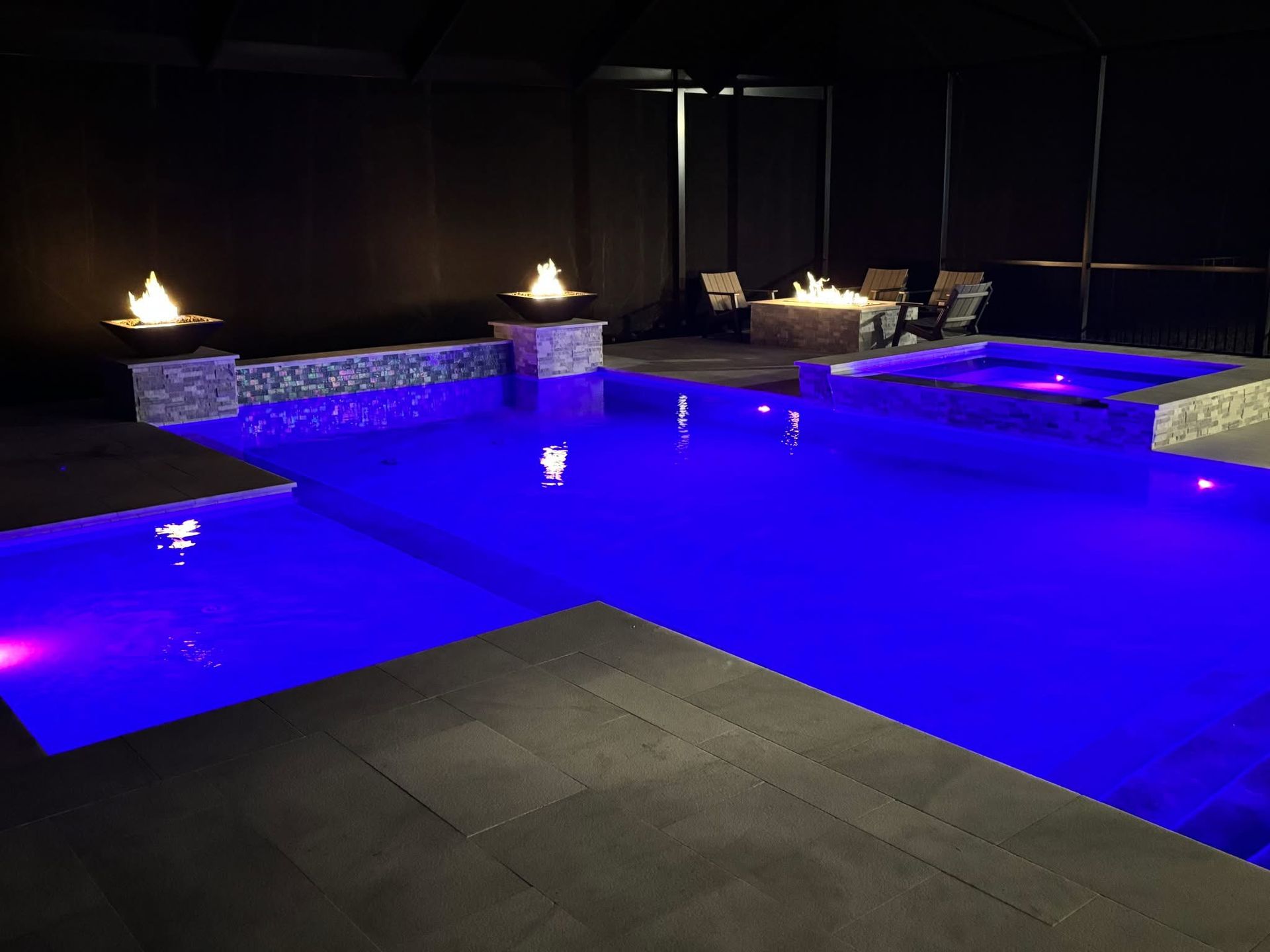 Nighttime view of a pool and hot tub with blue lights, fire bowls, and a small seating area.