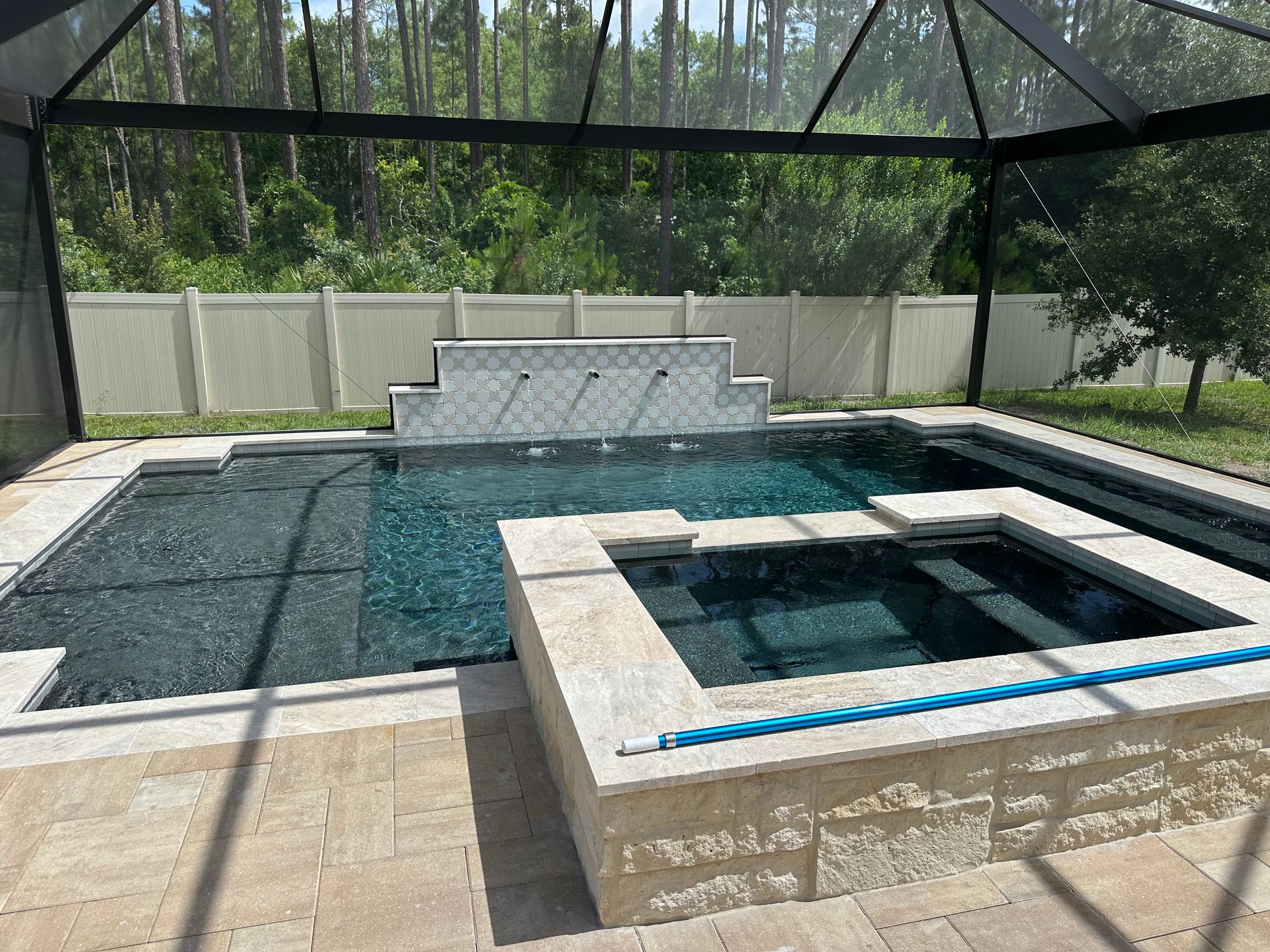 A backyard pool with a spa, fountains, and a screened enclosure. The pool is surrounded by a tan patio and a white fence.