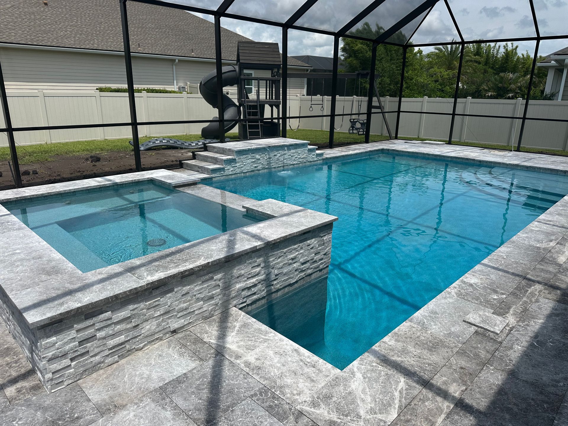 Swimming pool with attached spa, surrounded by a screened enclosure. The pool has blue water, gray tile, and a small slide.