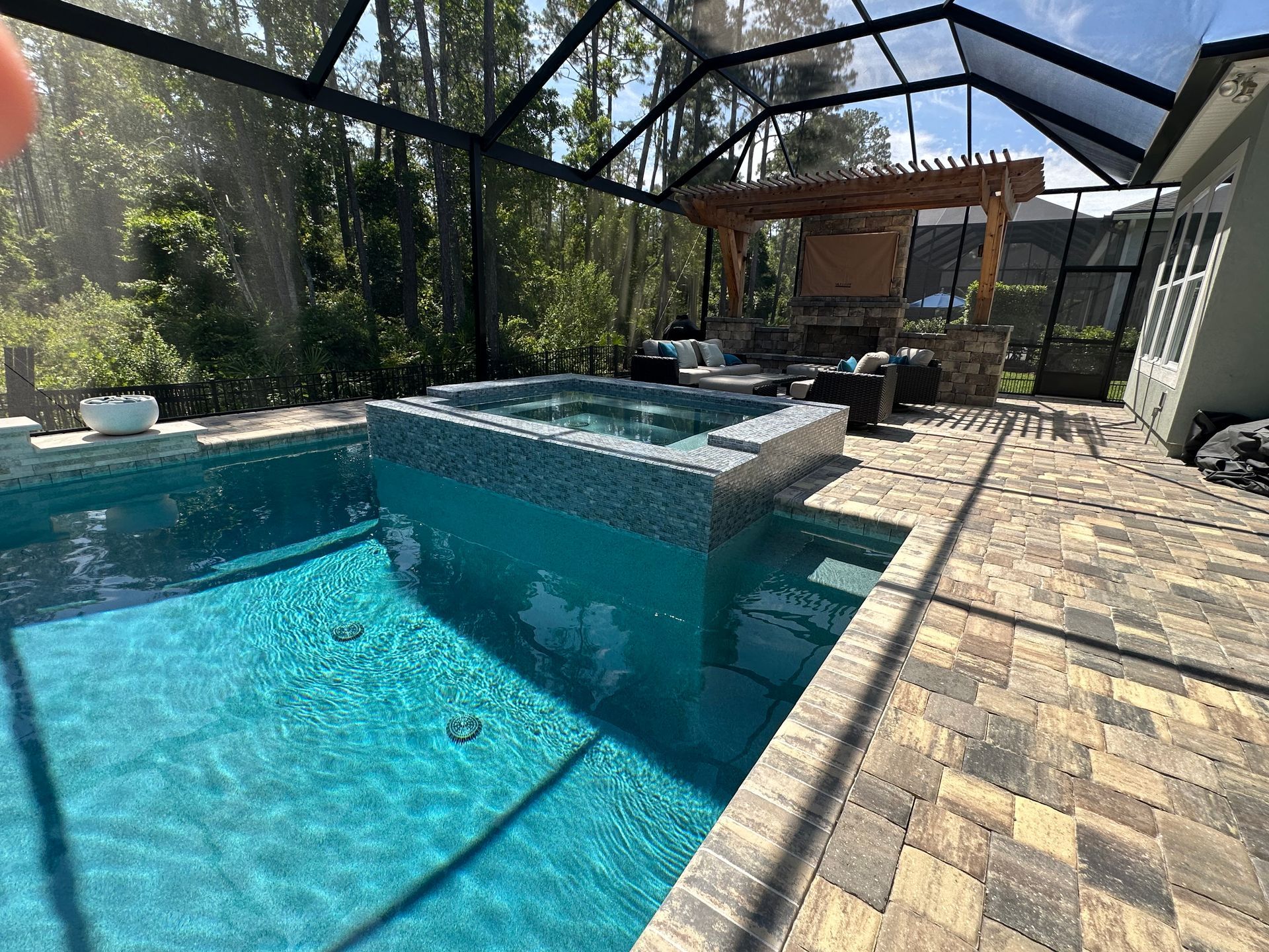 Pool area with a turquoise pool and hot tub, brown patio, and screened enclosure.  Outdoor kitchen and seating area in the background.