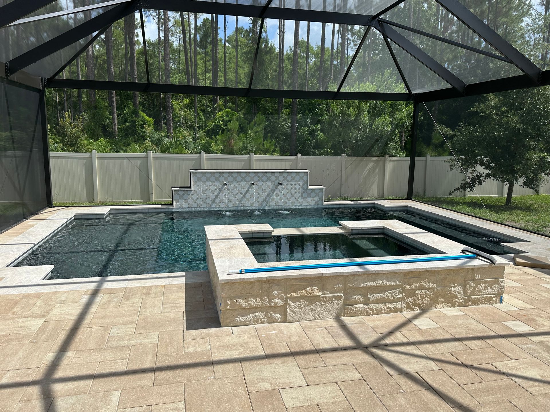 A backyard pool with a raised spa and waterfall feature, enclosed by a screen and white fence. Light-colored paving surrounds the pool.
