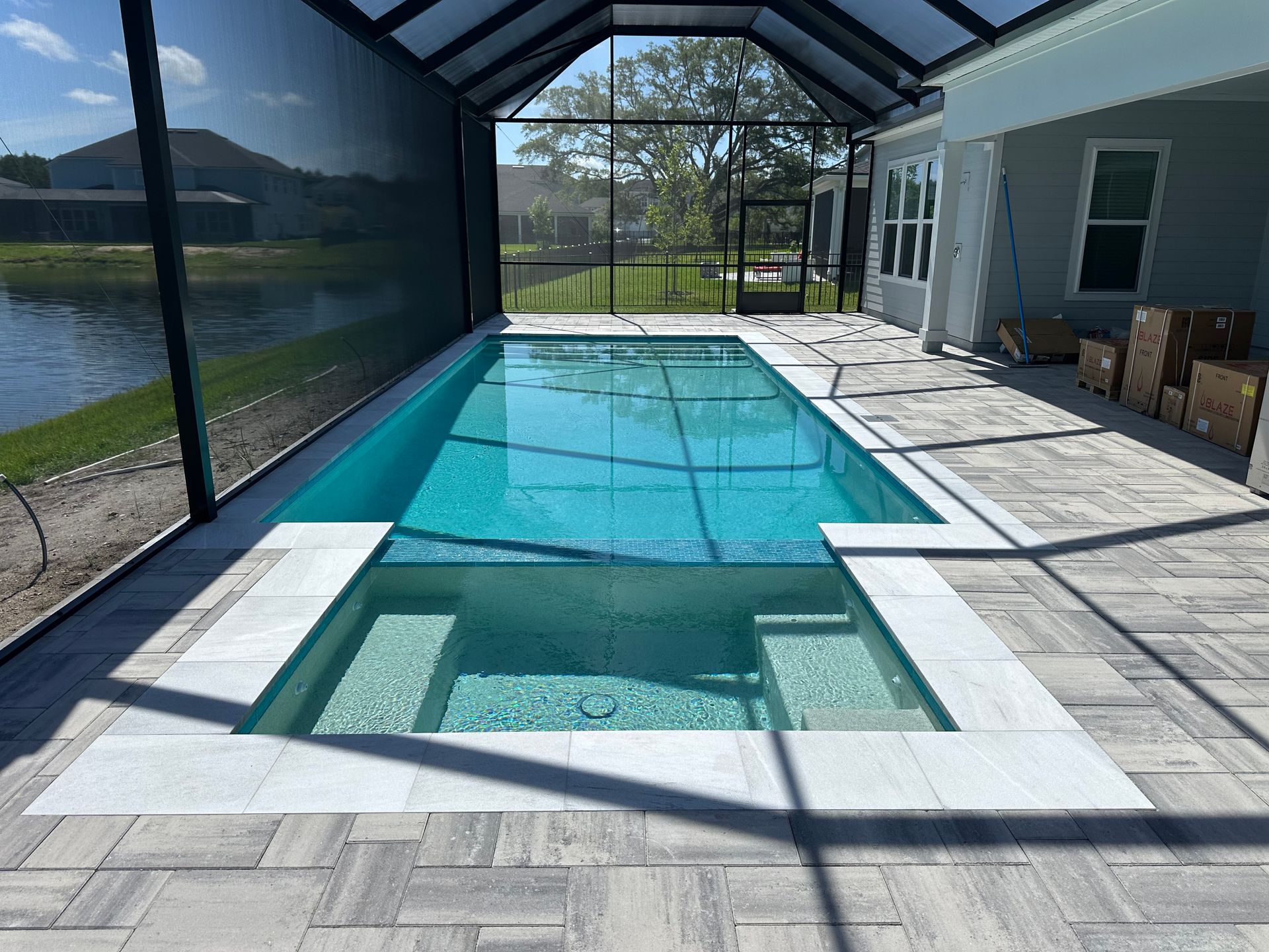 A rectangular pool and jacuzzi surrounded by light pavers, within a screened-in patio, overlooking a lake.