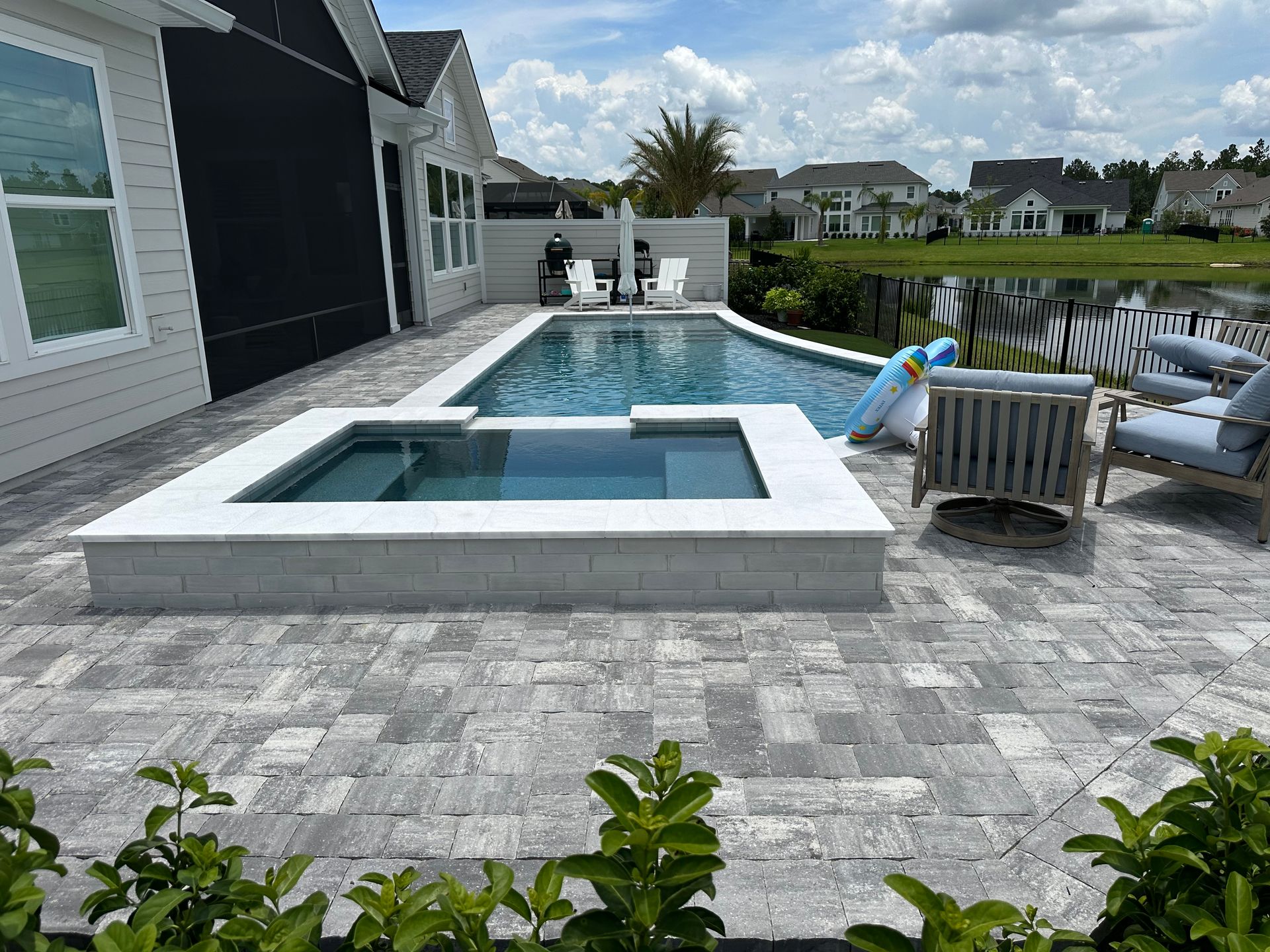 A backyard pool and spa with a brick patio, facing a lake. Lounge chairs and a floating inflatable are visible.