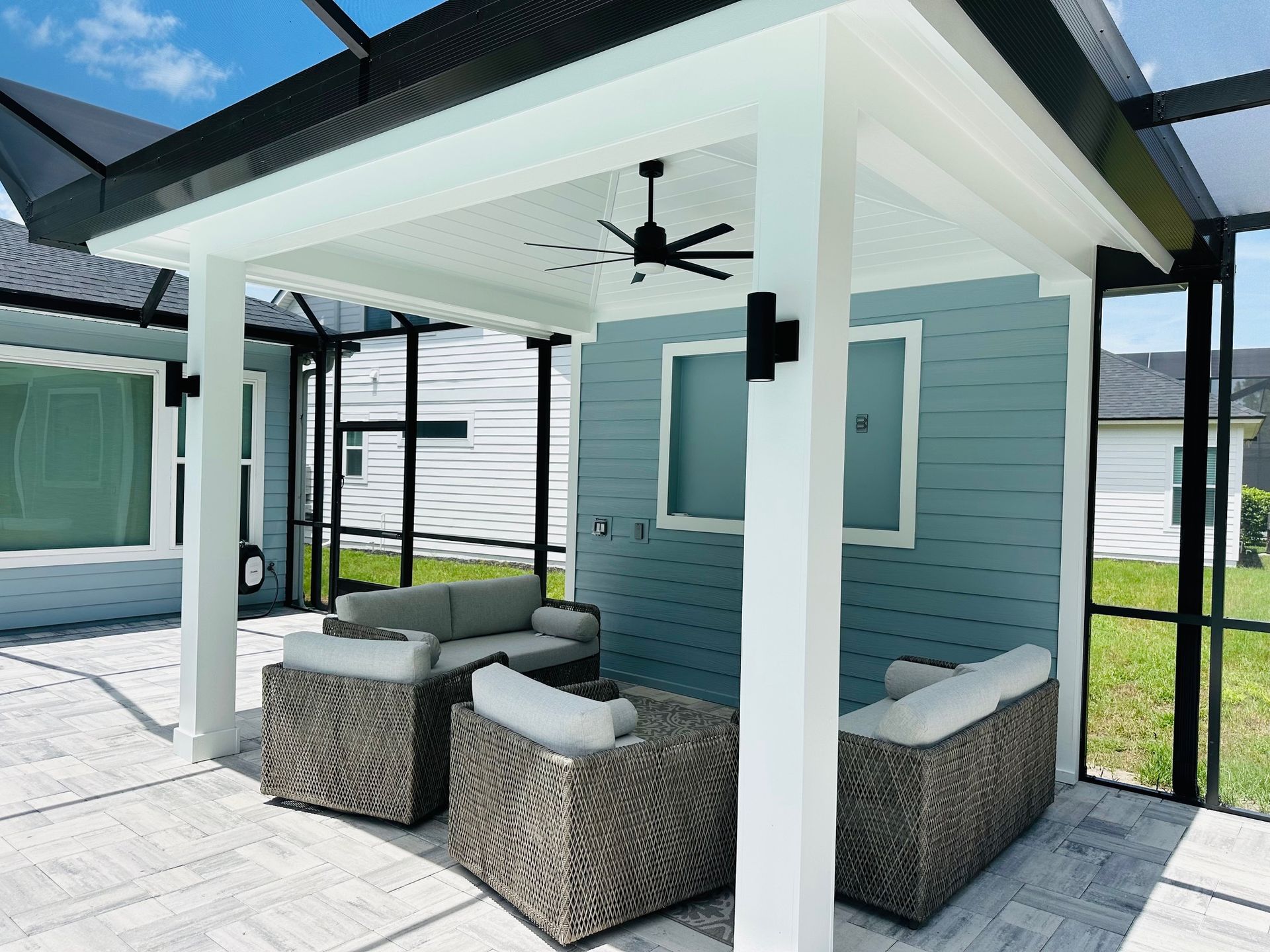 Outdoor patio with seating under a white pergola with a ceiling fan. Gray wicker furniture on a stone patio, blue wall backdrop.