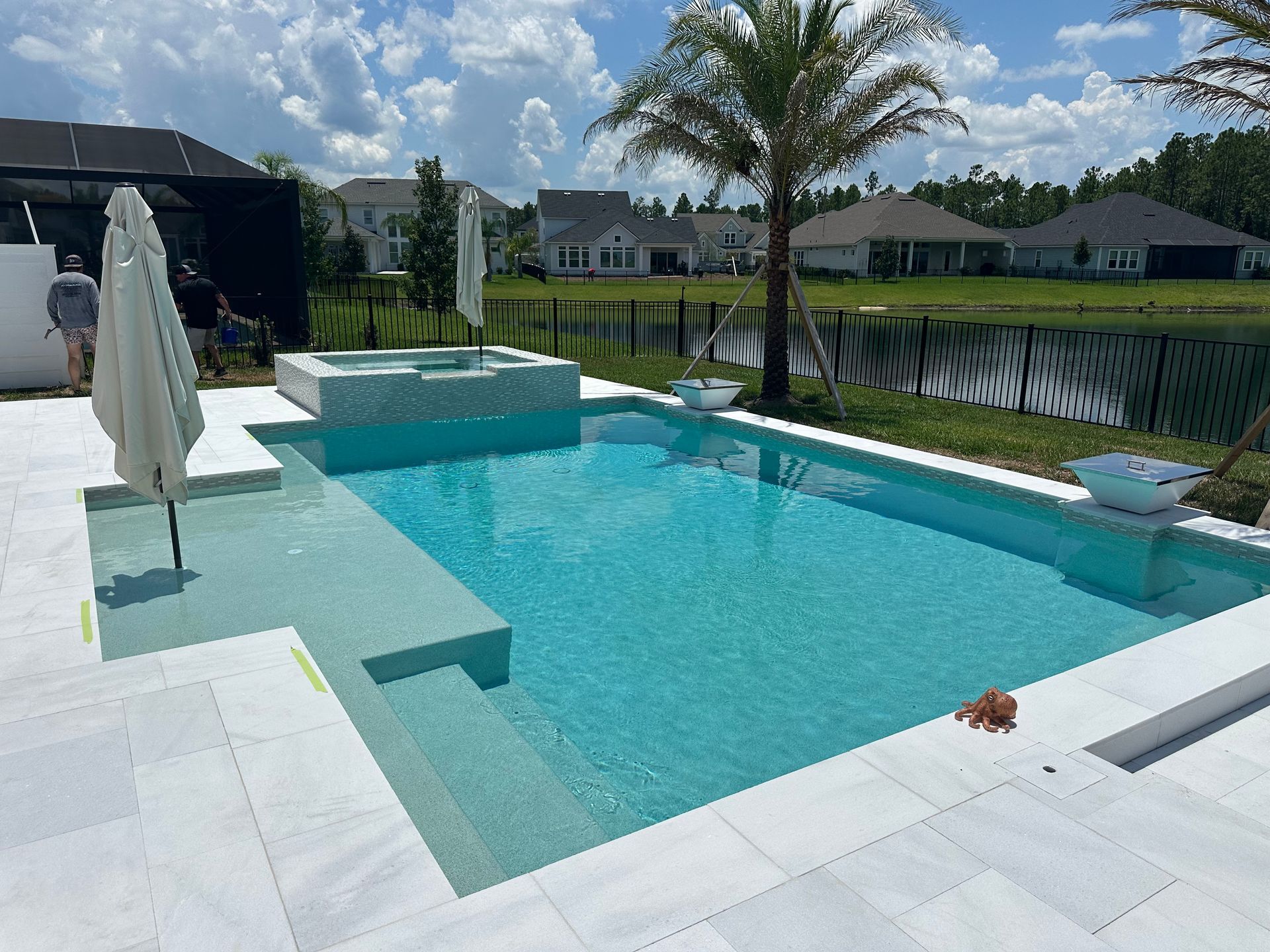 A bright turquoise pool with a raised spa area. The pool is surrounded by white tiling, with umbrellas, and a view of houses and a lake in the background.