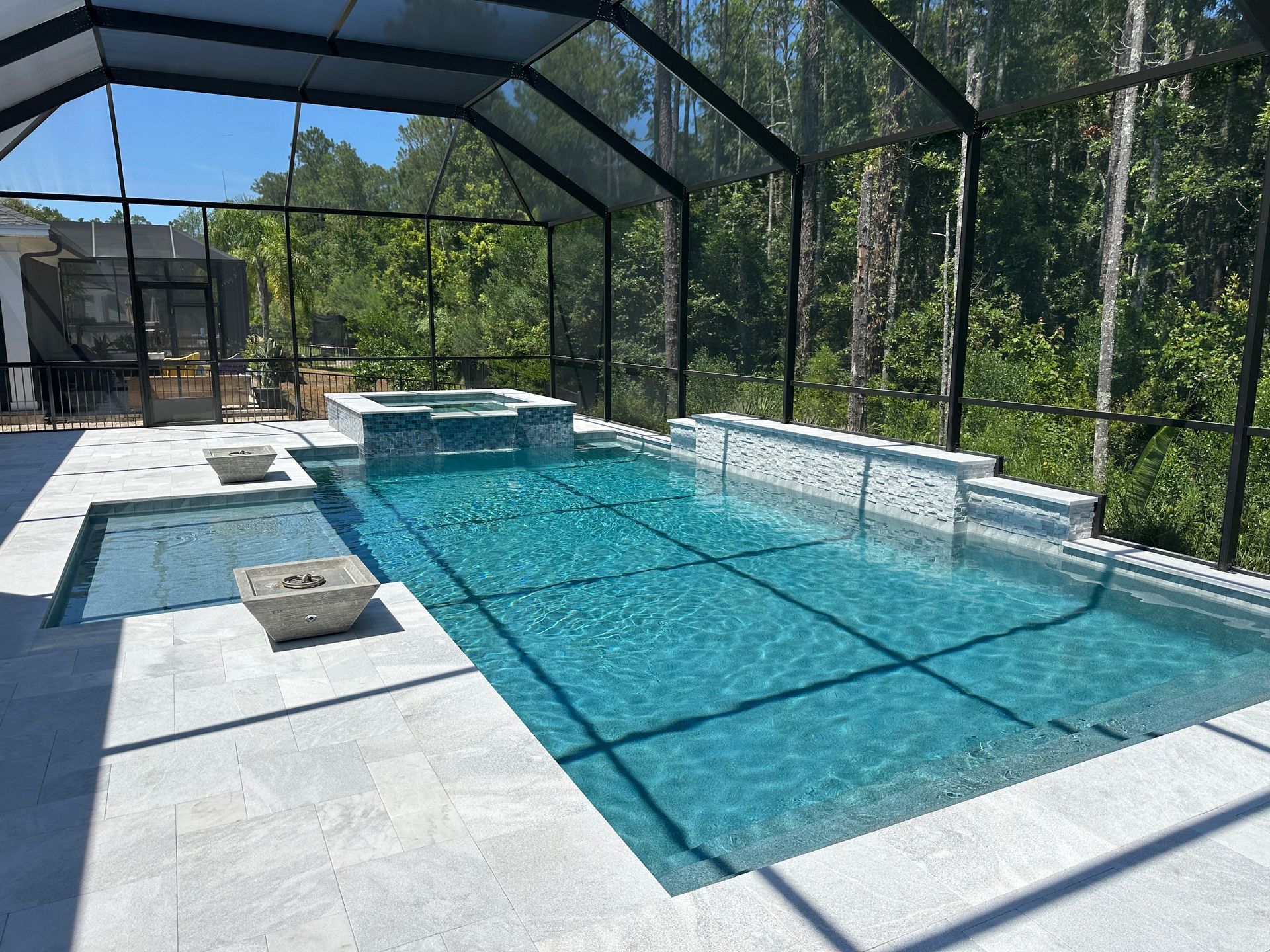 A luxurious outdoor pool and spa area with a screen enclosure, surrounded by a stone patio and lush greenery.