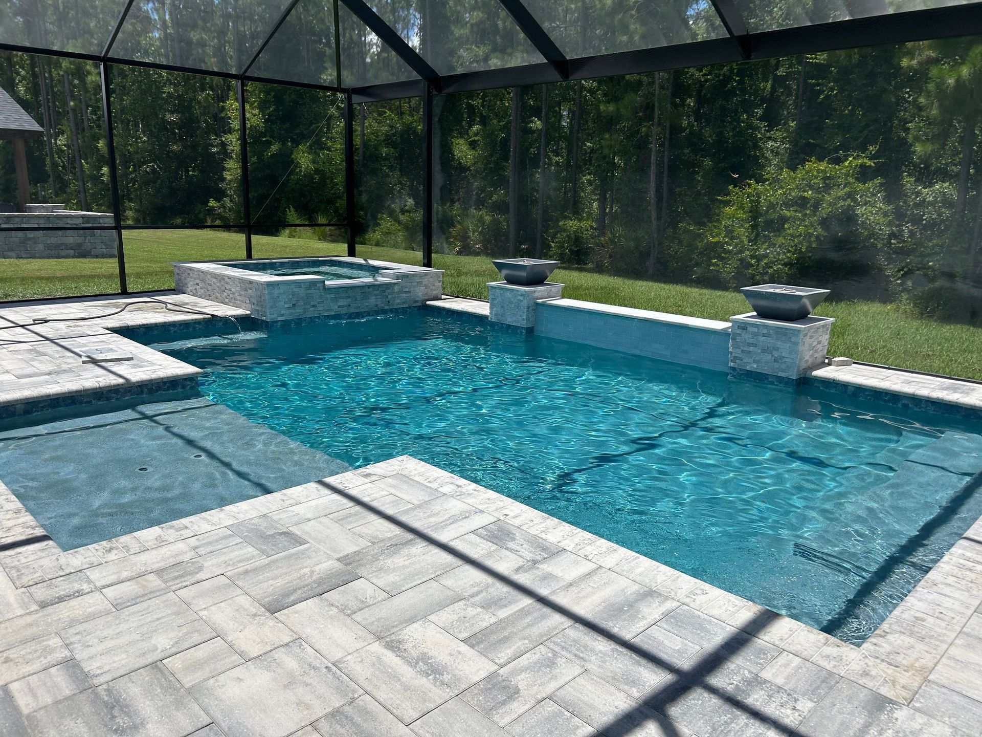 A rectangular pool and spa enclosed by a screen, surrounded by gray paving stones. The water is turquoise, and lush greenery is in the background.