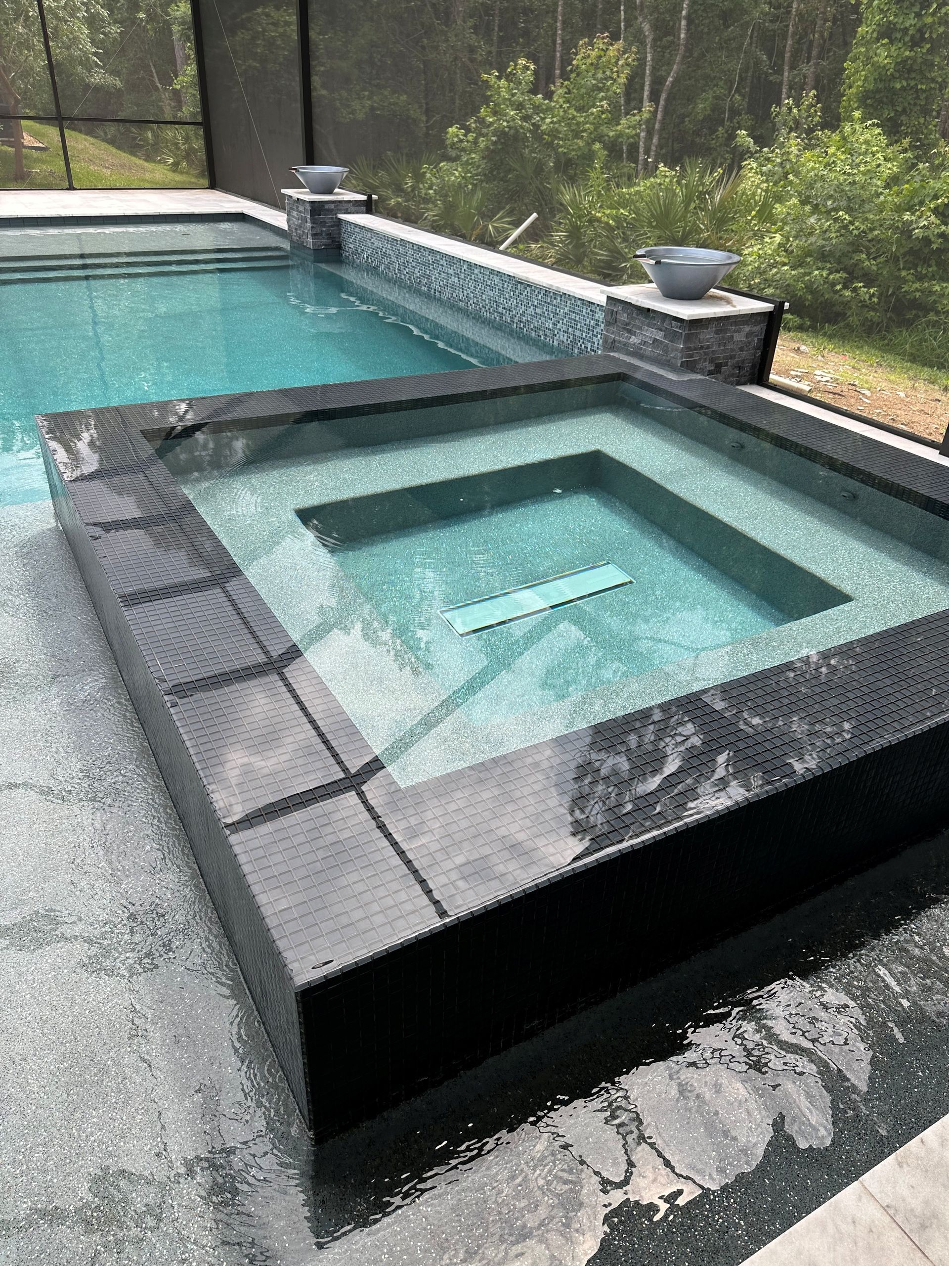 A square spa with black tile is next to a pool. Both are filled with blue-green water. The setting is outdoors.