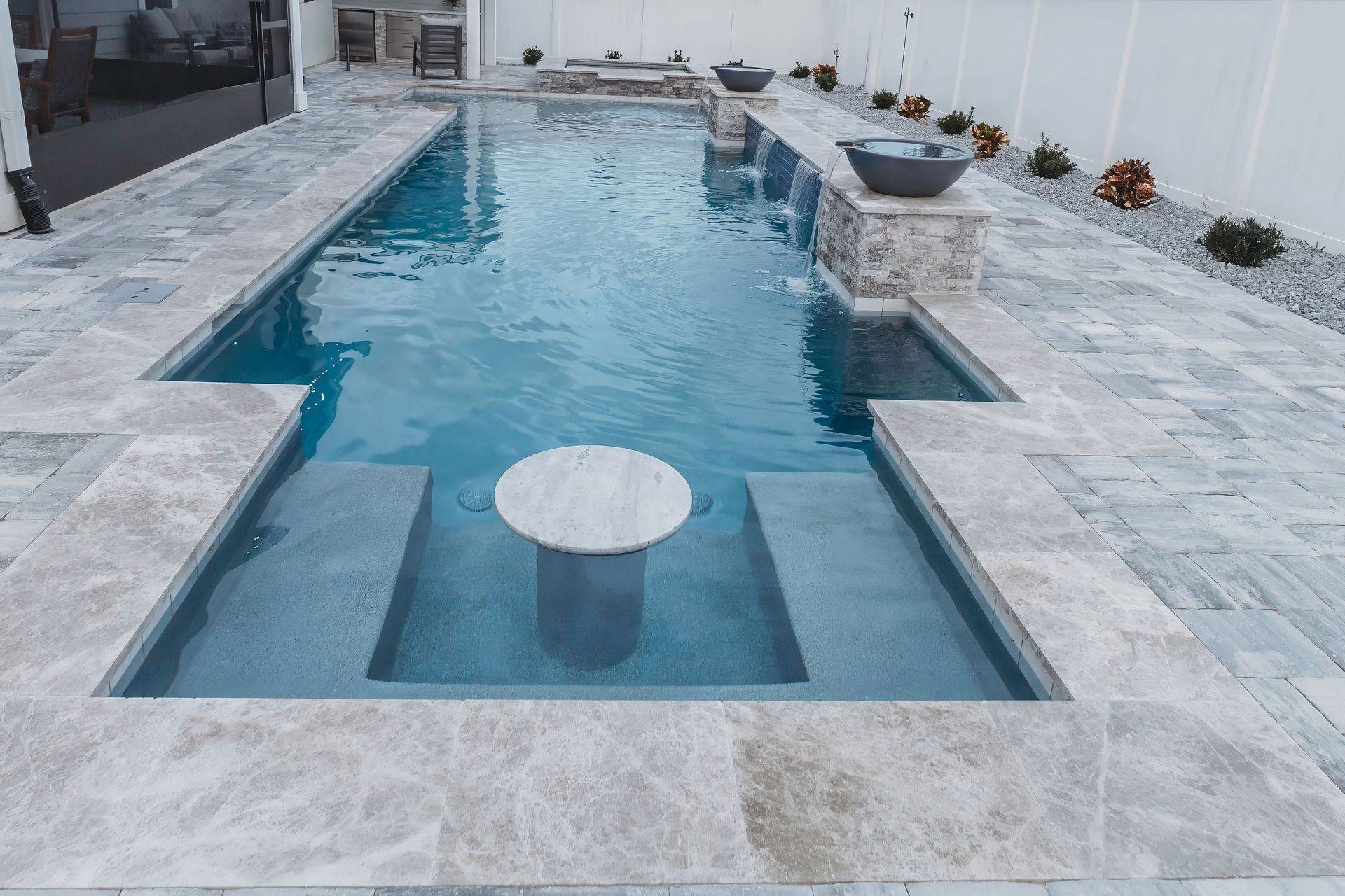 Pool with a central table, stone tile surrounding the pool, and a water feature flowing from a stonework wall.