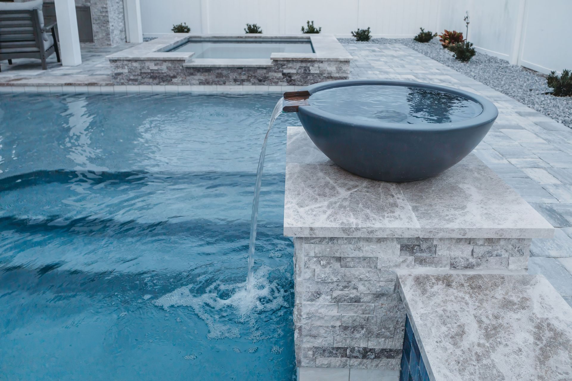 A gray concrete water feature spills water into a pool. The pool has a stone patio and hot tub in the background.
