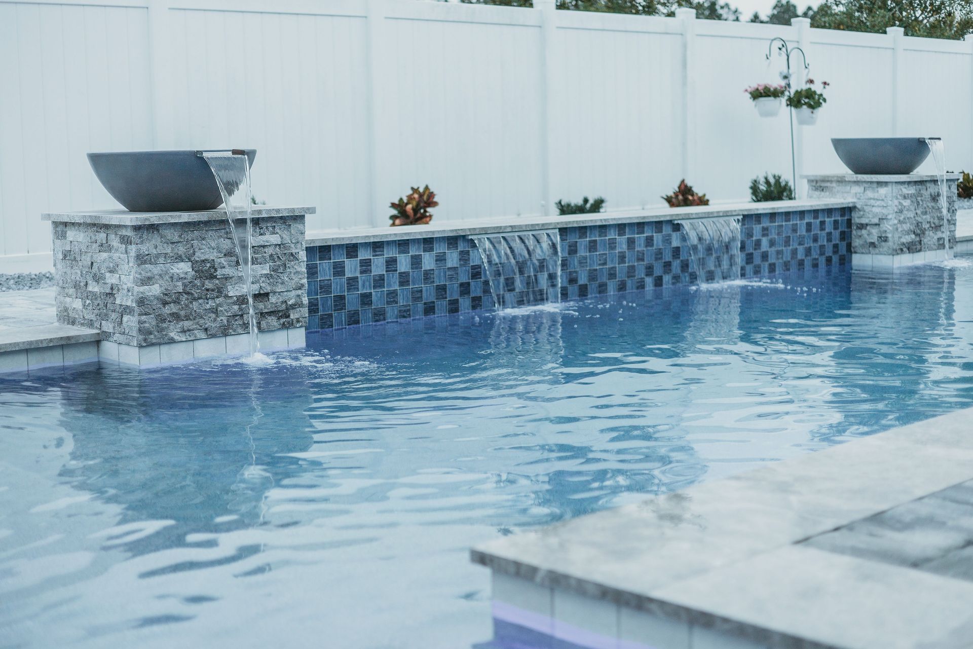 Pool with a waterfall feature; water flows from blue-tiled wall. Planters and fountains on stone structures, white fence in background.