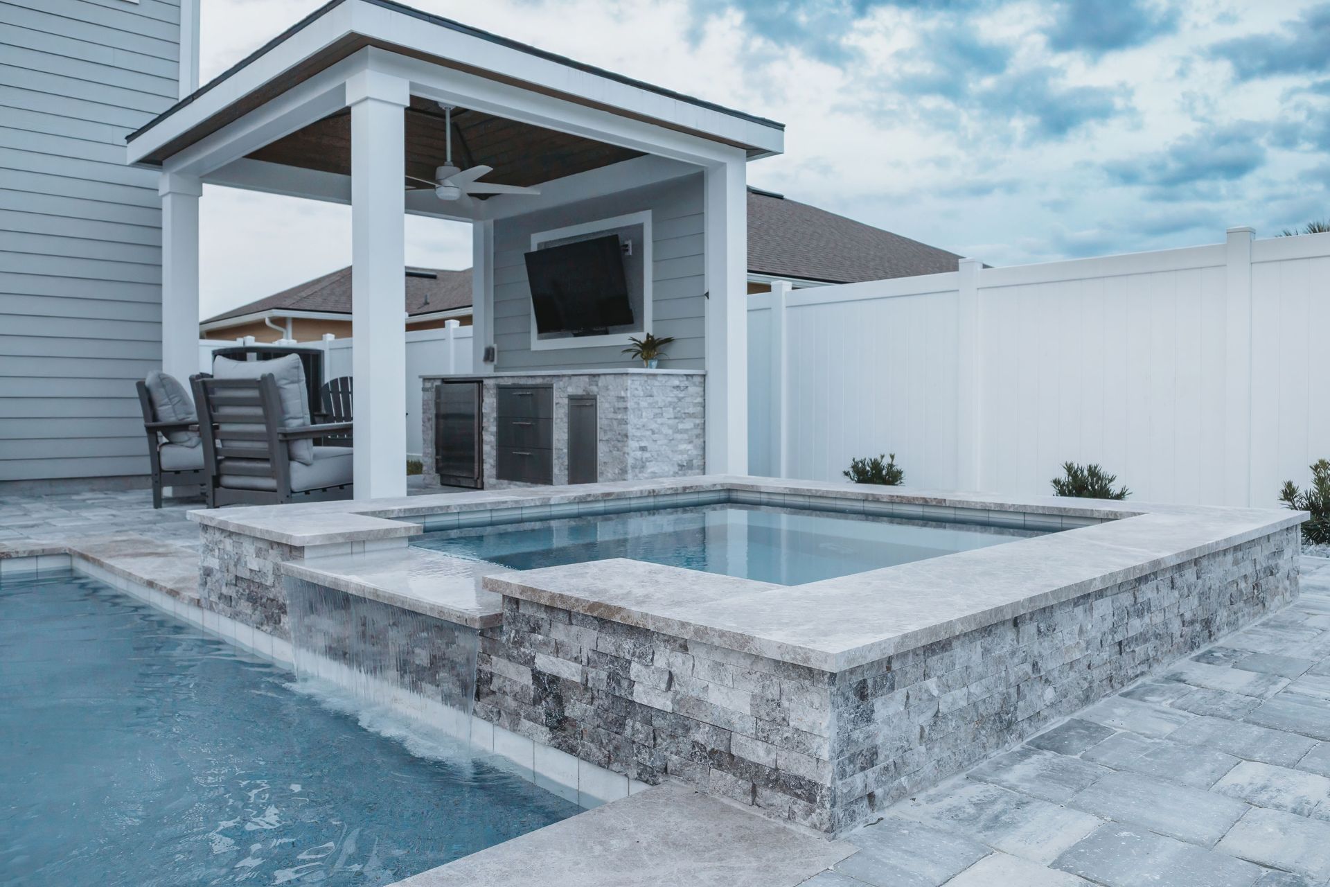 Outdoor pool and spa area with a small structure featuring a TV, bar, and seating. Light blue pool water, grey stone accents, and white fence.
