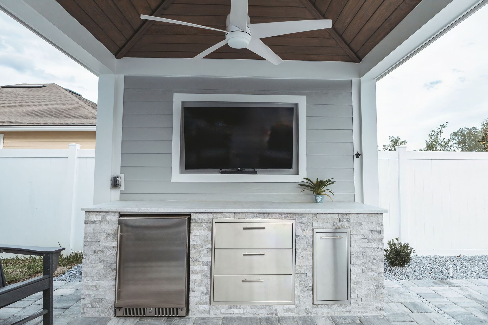 Outdoor kitchen with gray siding, a mounted TV, and stainless steel appliances, under a white pergola with a ceiling fan.