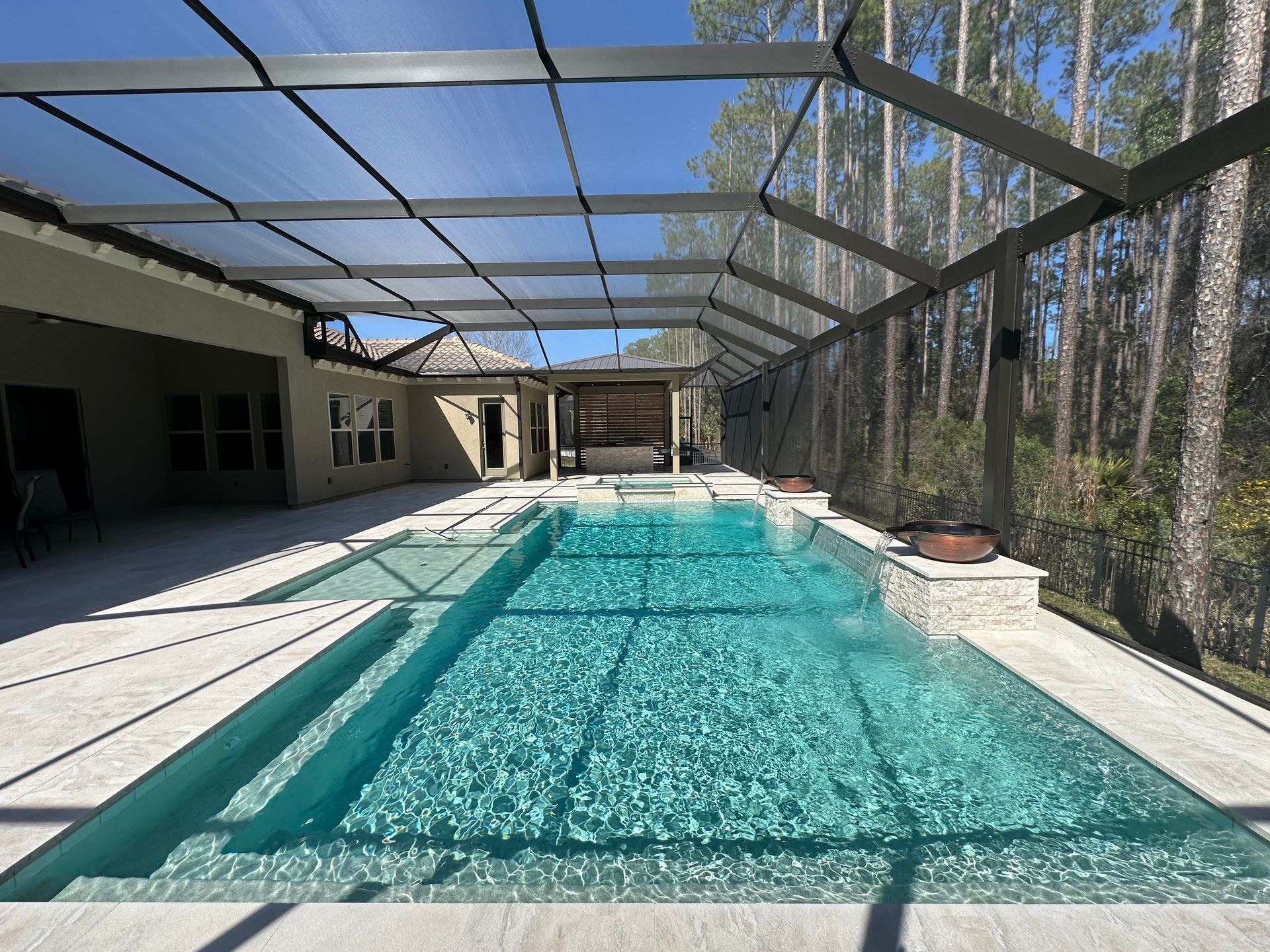 A rectangular swimming pool under a screened enclosure, next to a patio and lush trees. Bright blue water reflects sunlight.