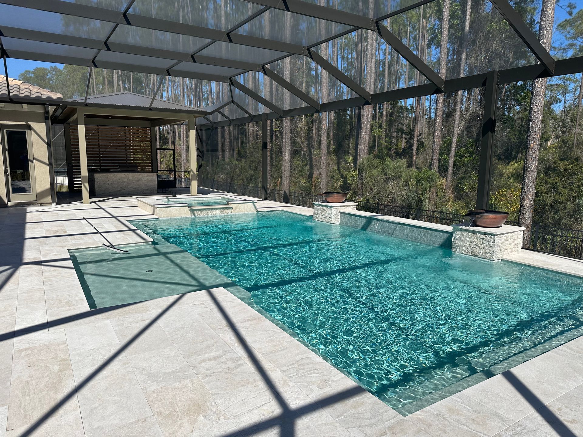 A covered outdoor pool with turquoise water, surrounded by light-colored stone and landscaping. A forest is visible in the background.