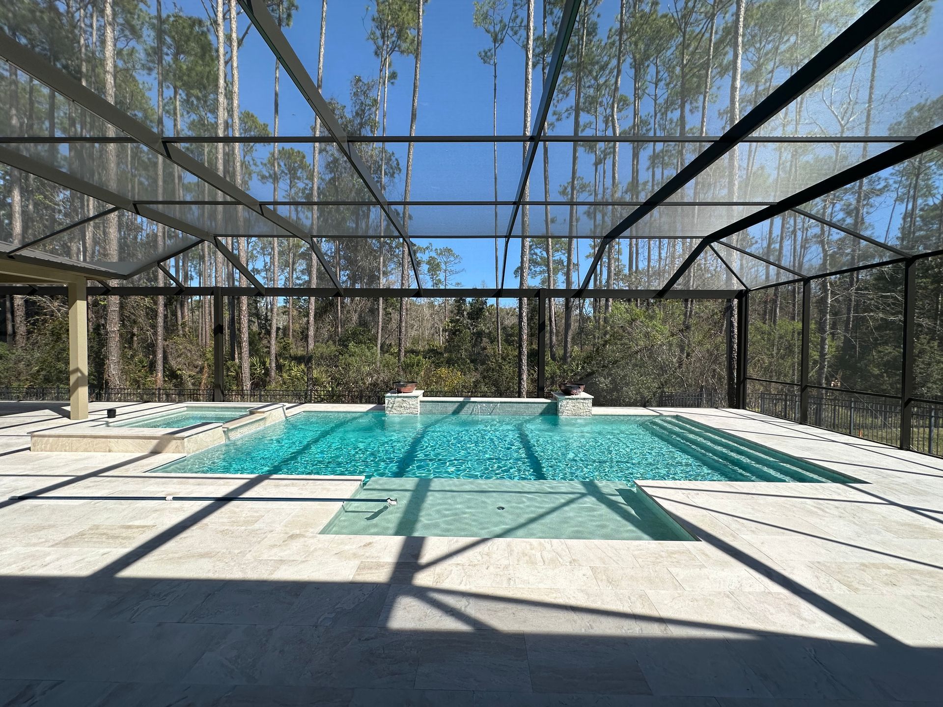 Swimming pool under a screened enclosure with a forest background. Blue water, light stone patio, and a clear, sunny sky.