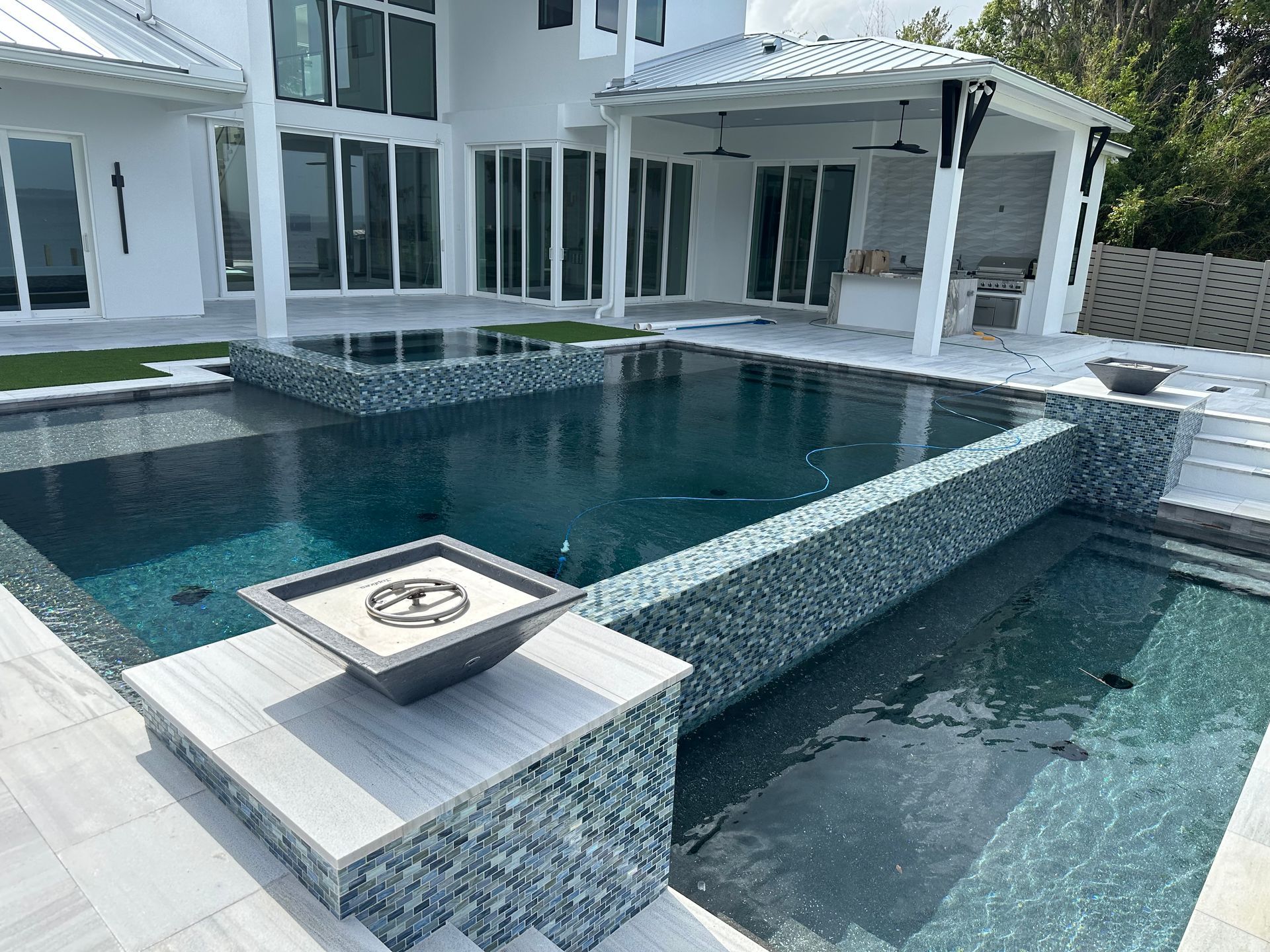 Modern luxury backyard with a pool, spa, and outdoor kitchen.  White house with a covered patio, dark blue water, and gray and blue tiling.
